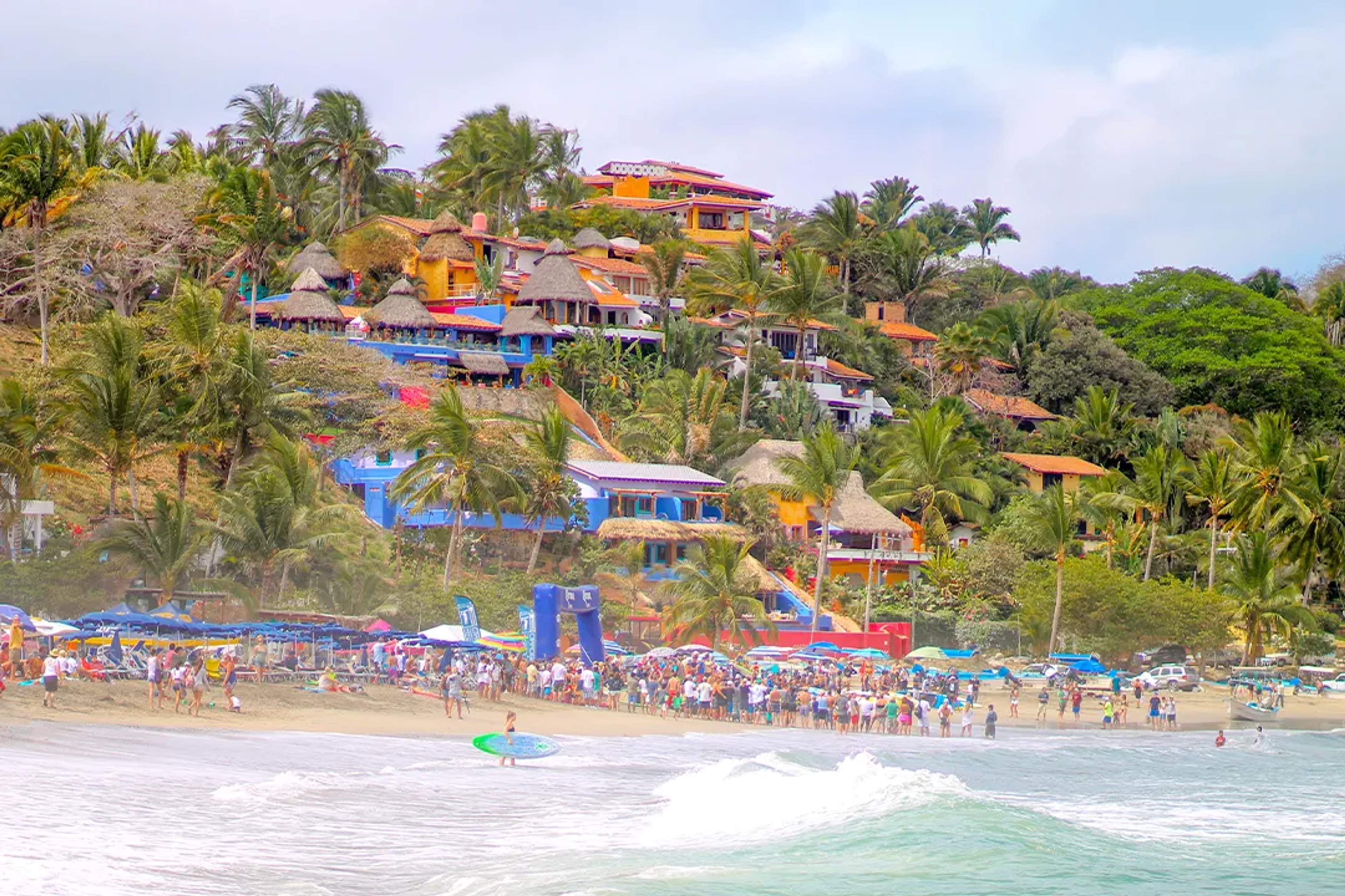 Colorful hillside homes and a lively beach scene in Sayulita, filled with people and surf vibes.