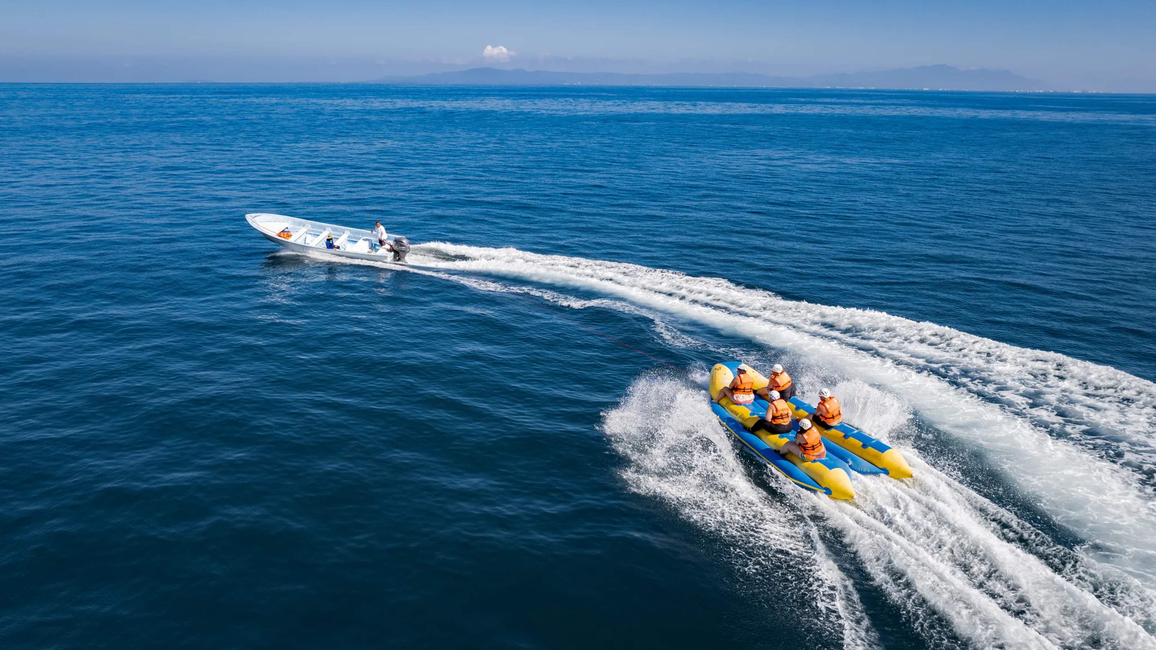 Yellow inflatable banana boat with passengers bouncing over ocean waves, support boat following close behind in clear blue