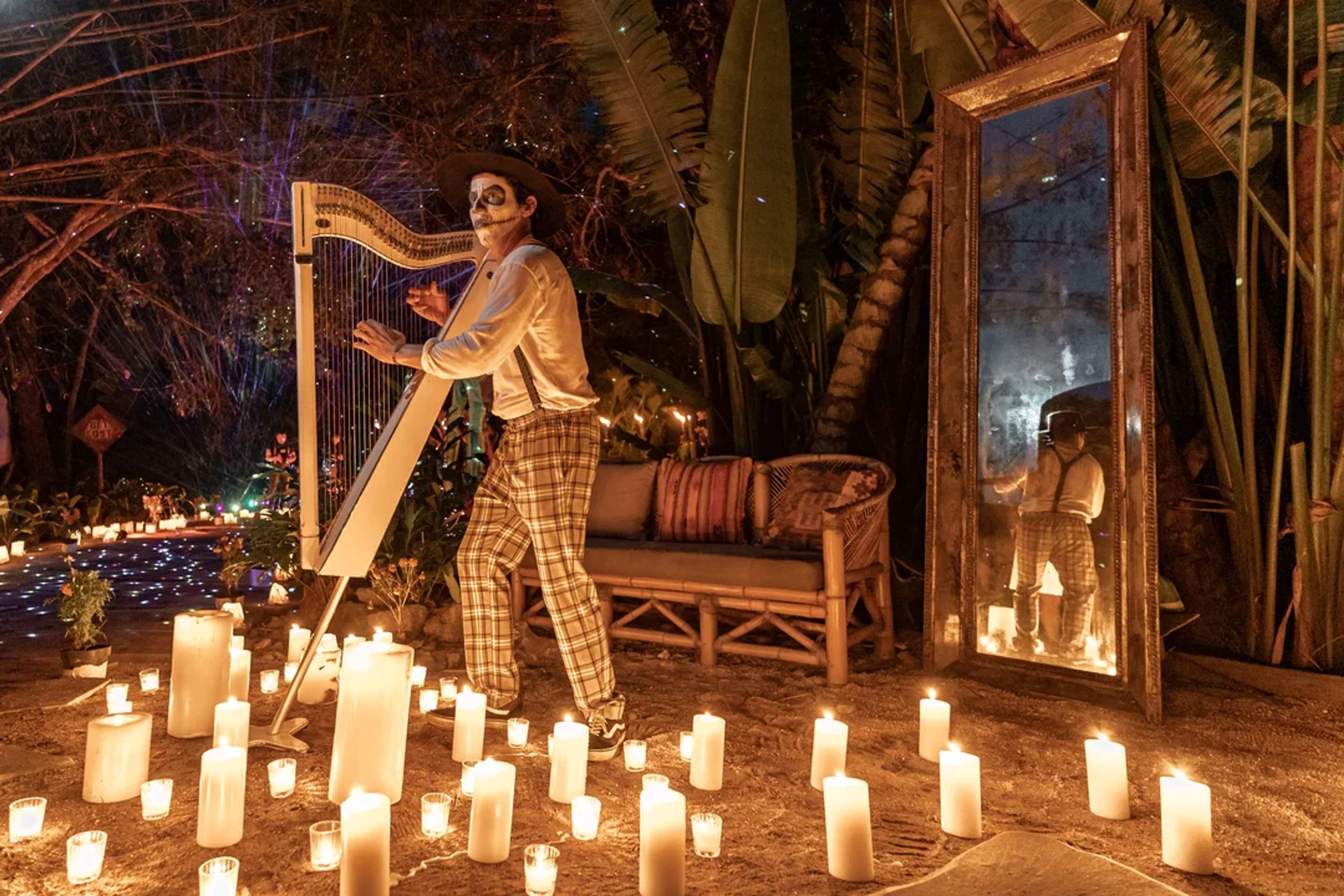 Musician with face paint plays harp among candles during a Día de Muertos celebration at night.