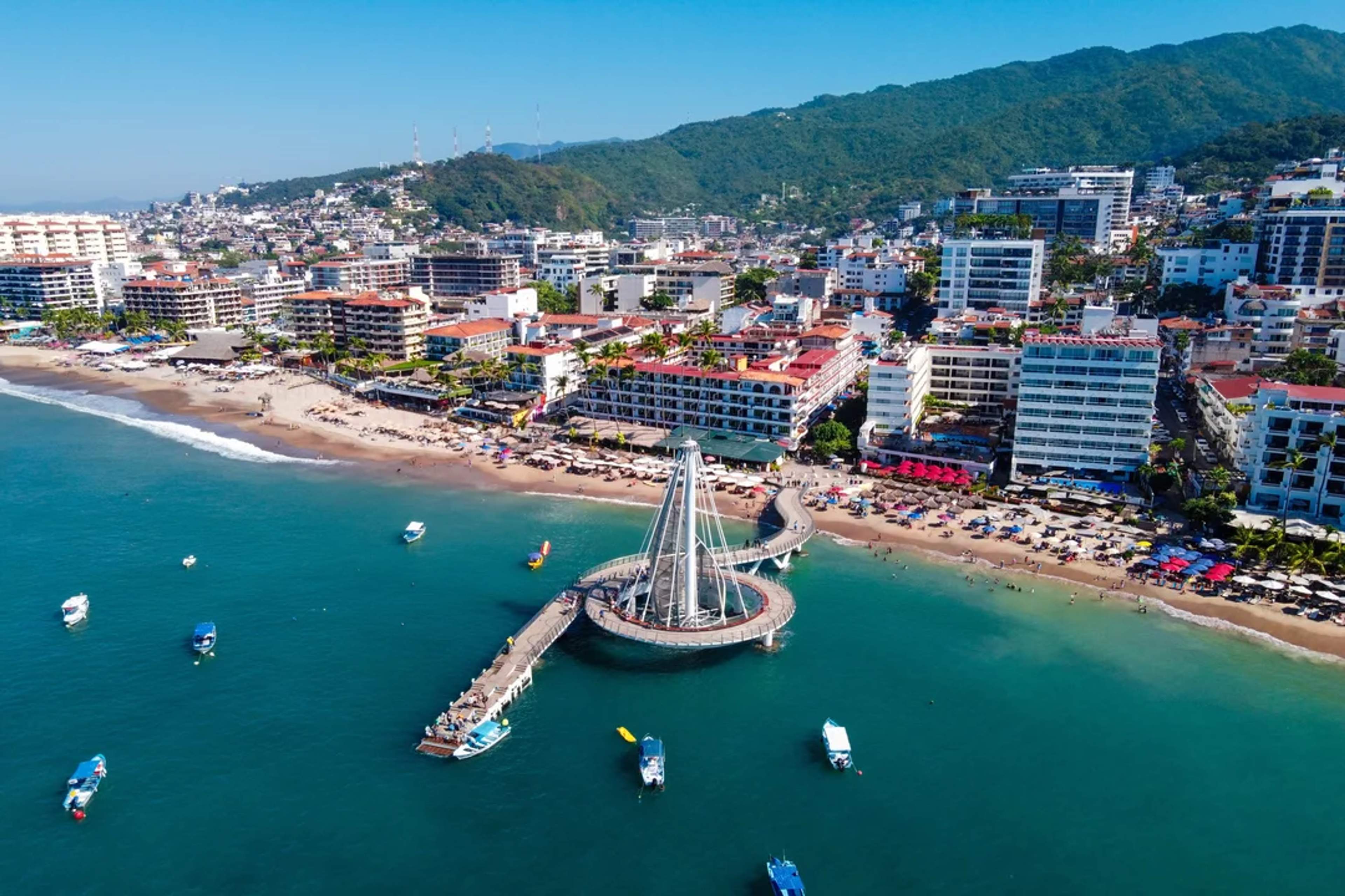 Vista aérea del muelle de Puerto Vallarta, su playa vibrante y montañas verdes al fondo.