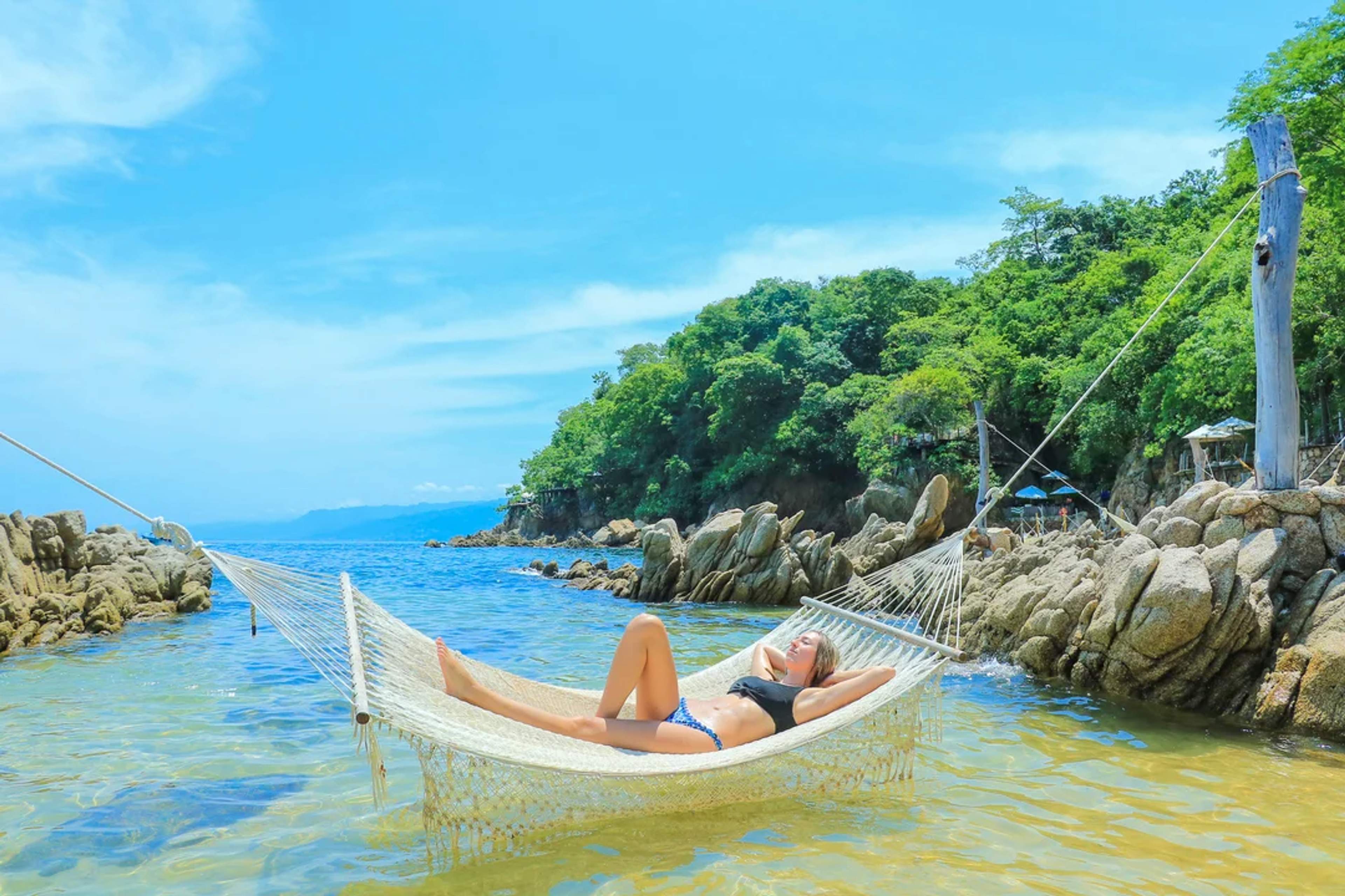 Woman relaxing on a hammock over clear water, surrounded by rocks and lush tropical forest.