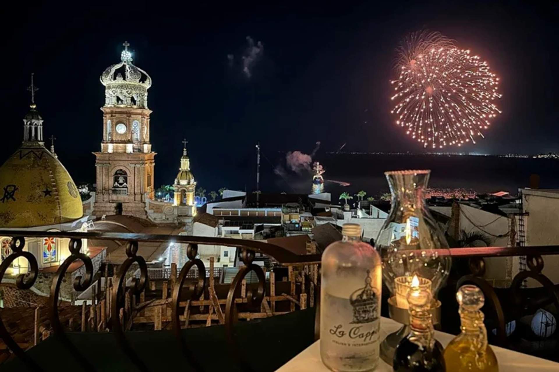 A night view of Puerto Vallarta with fireworks lighting up the sky above the city. The foreground features a table with a bottle, glasses, and candles, set on a balcony overlooking the illuminated church towers and rooftops.