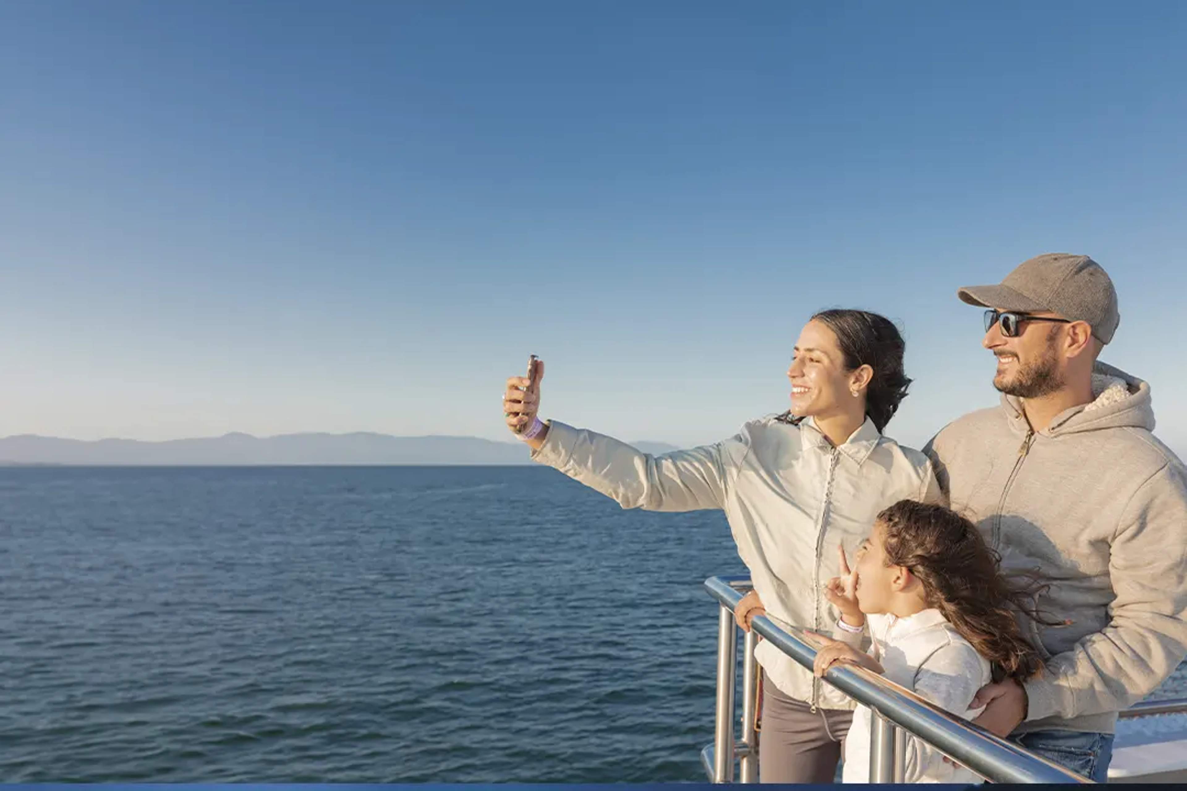A family taking a selfie on a boat, with the ocean and distant mountains in the background under a clear blue sky.