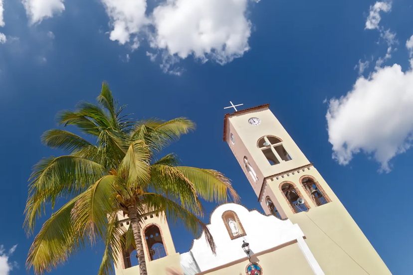 Una palmera junto a una iglesia beige con torre de reloj y cruz, contra un cielo azul brillante con nubes dispersas.