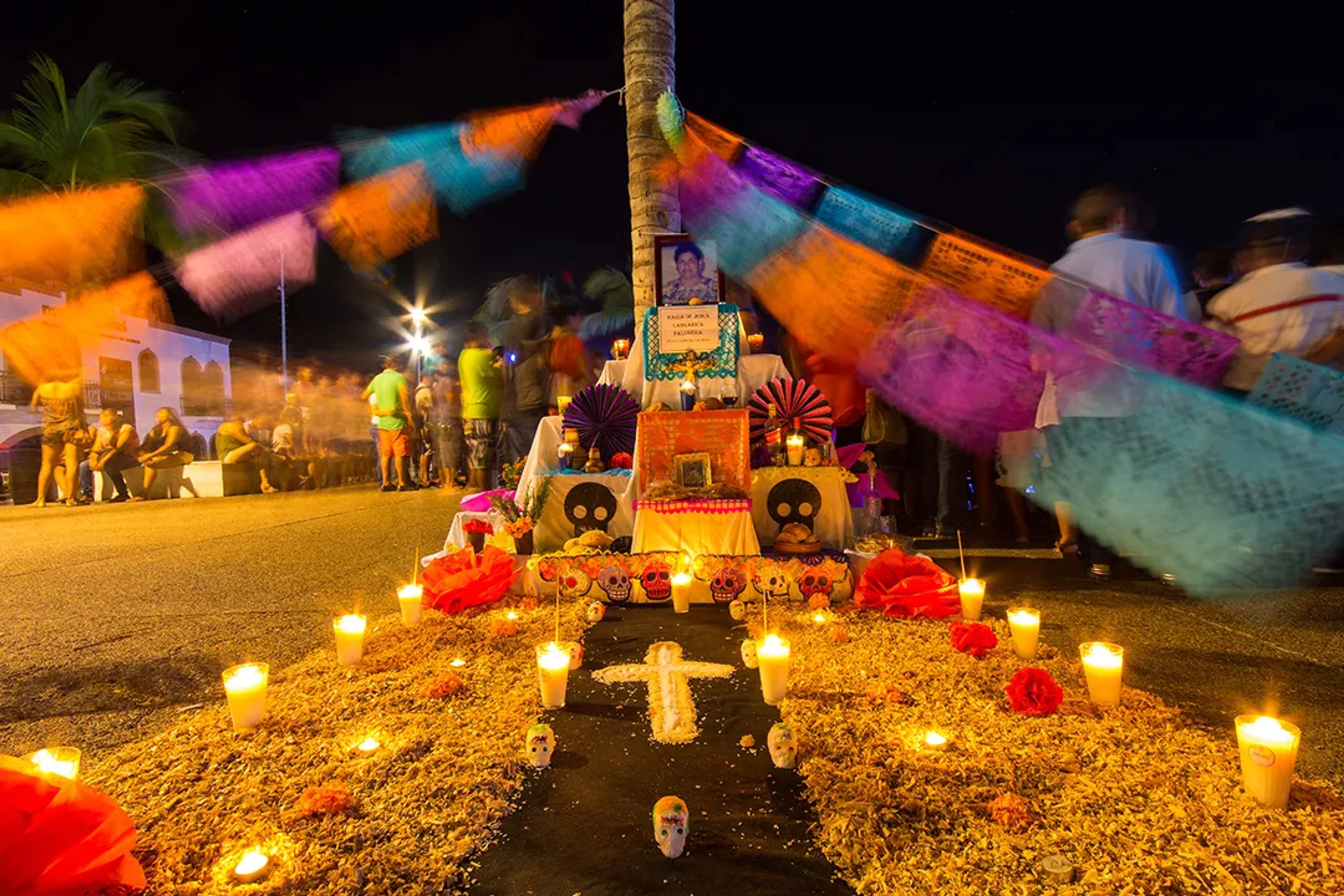 Ofrenda de Día de Muertos en la calle con velas, papel picado, cempasúchil y calaveras coloridas.