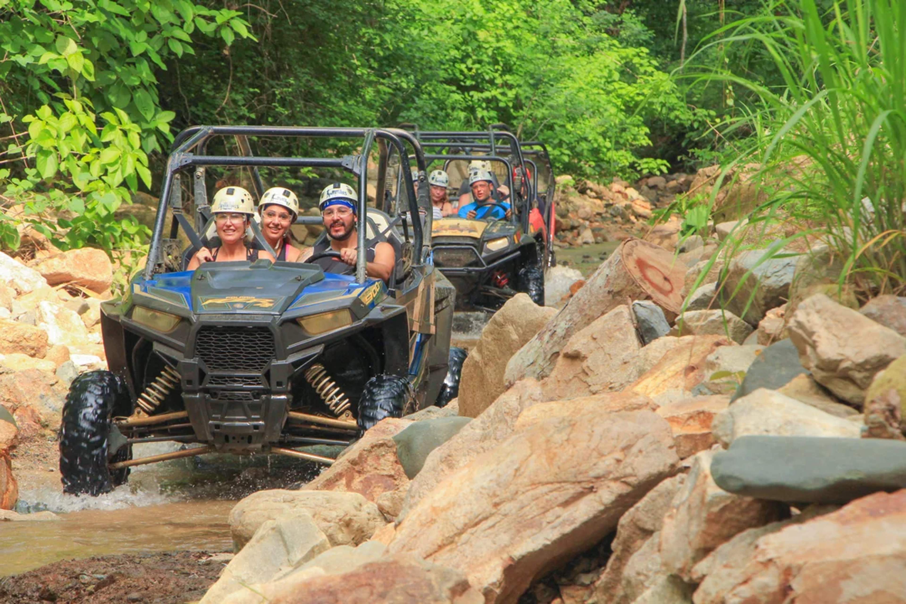 A group rides all-terrain vehicles through a rocky jungle trail, splashing through water and enjoying the off-road adventure.