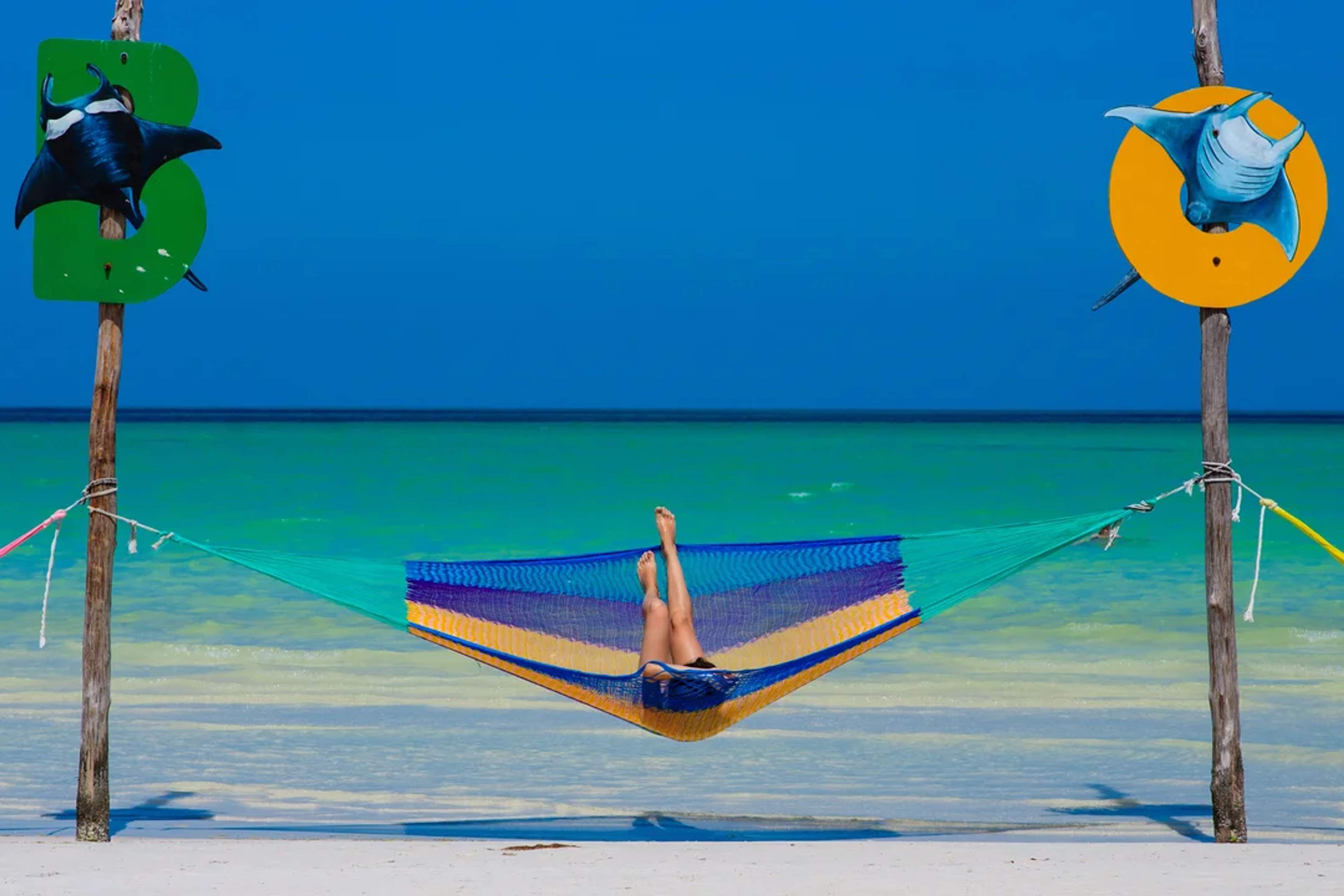 A person relaxes in a colorful hammock over the turquoise waters of a tropical beach under a blue sky.
