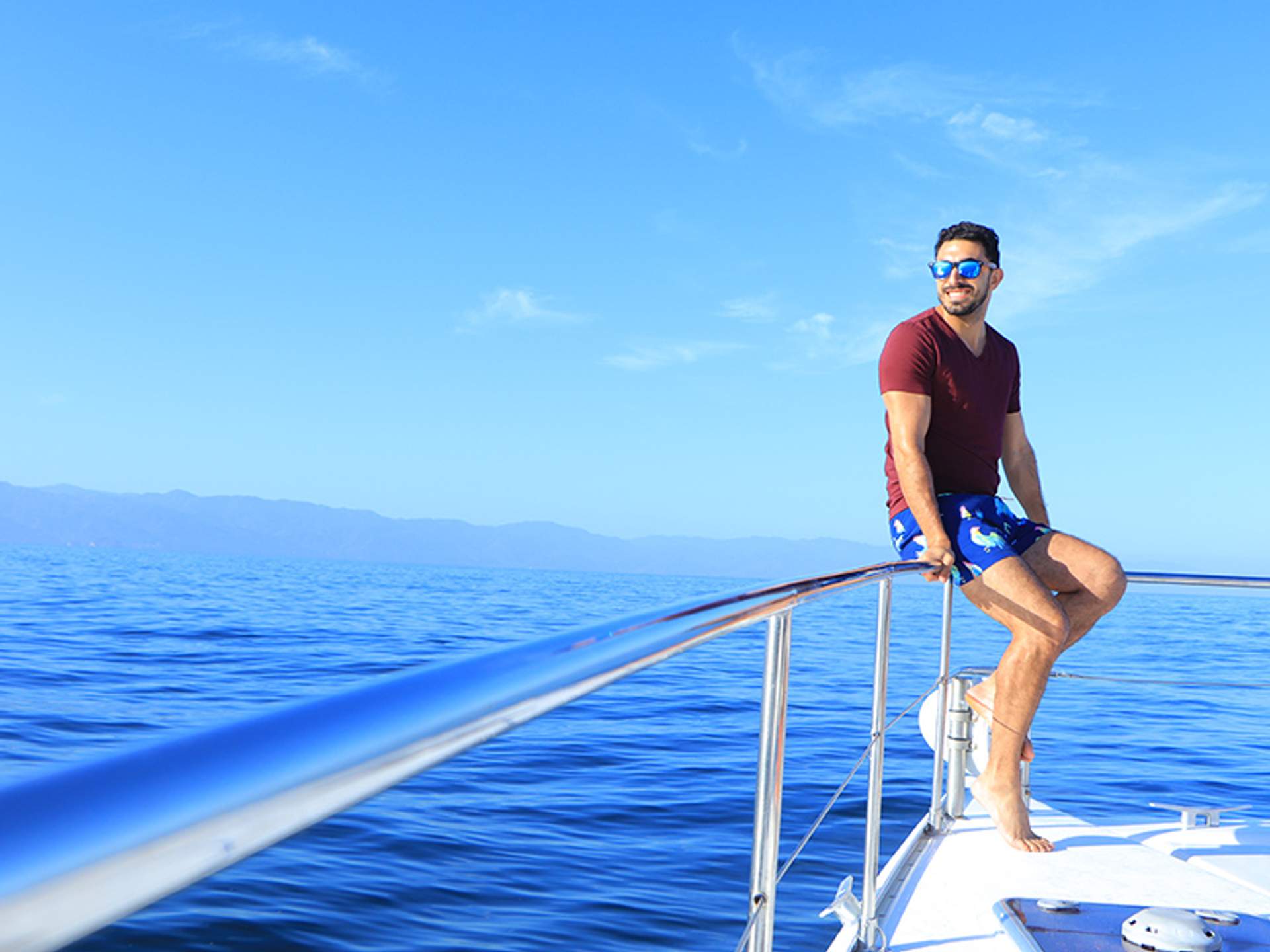 A man in sunglasses and swim trunks is sitting on the bow of a boat, smiling, with a vast blue ocean and distant mountains in the background.