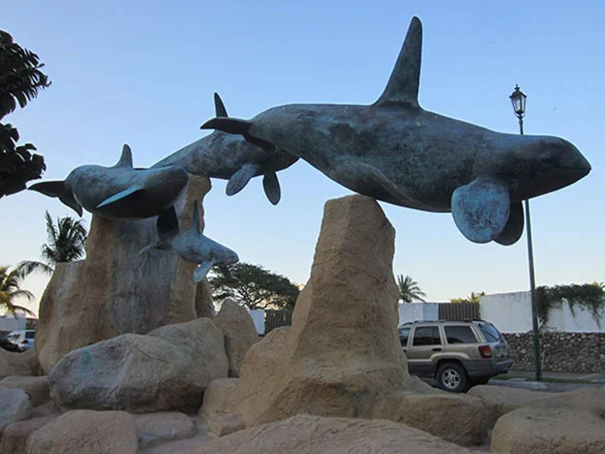 Escultura de orcas en vuelo sobre rocas en Puerto Vallarta, con un auto estacionado y palmeras al fondo.