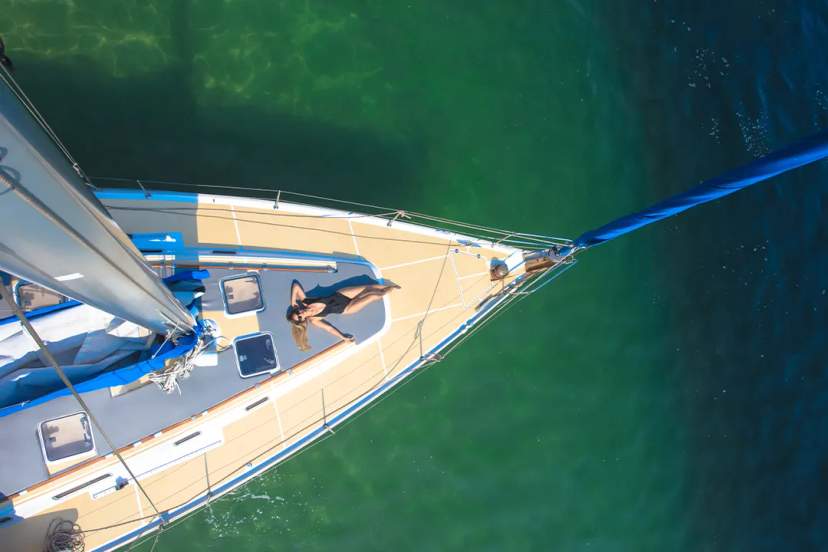 Woman sunbathes on the bow of a sailboat, floating over crystal-clear emerald waters.