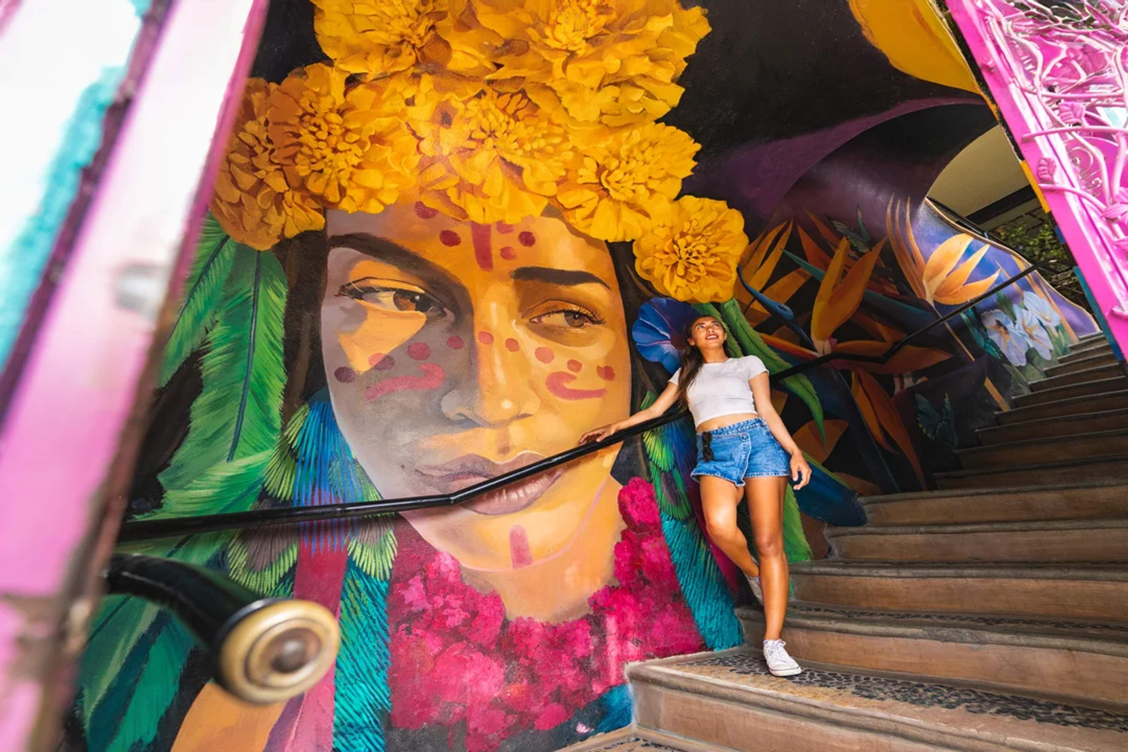 Colorful mural of a woman with marigolds, admired by a smiling girl on vibrant staircase.
