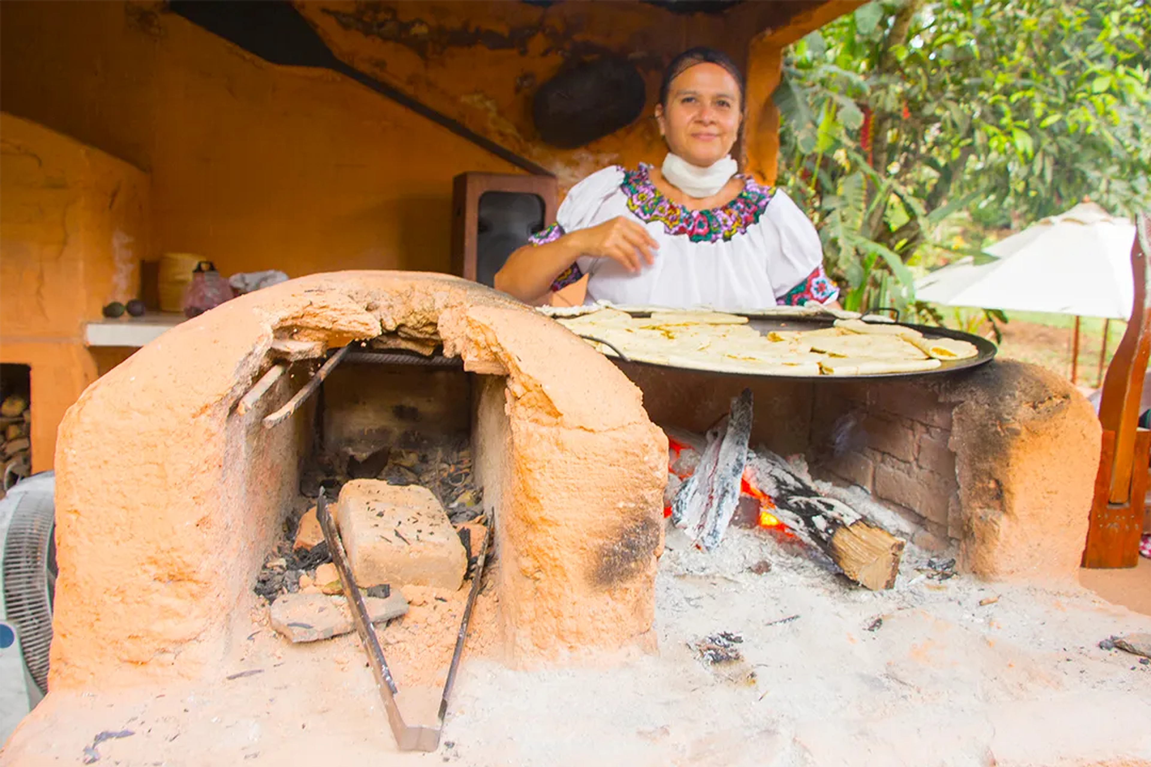 Woman in traditional dress cooking tortillas on a clay oven with wood fire in a rural setting.