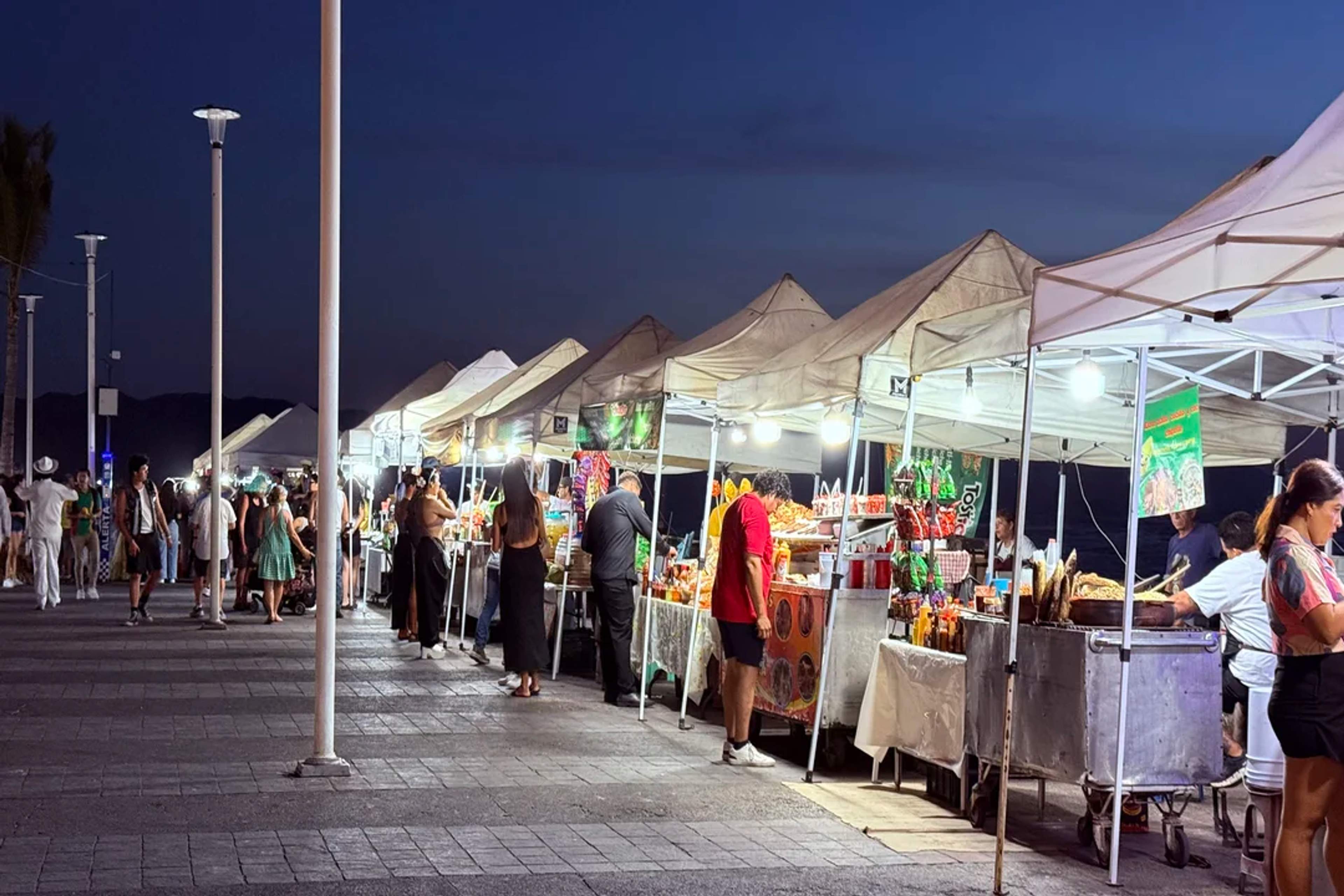 Cafetería acogedora de noche con luces de colores, sombrillas y gente disfrutando y conversando.