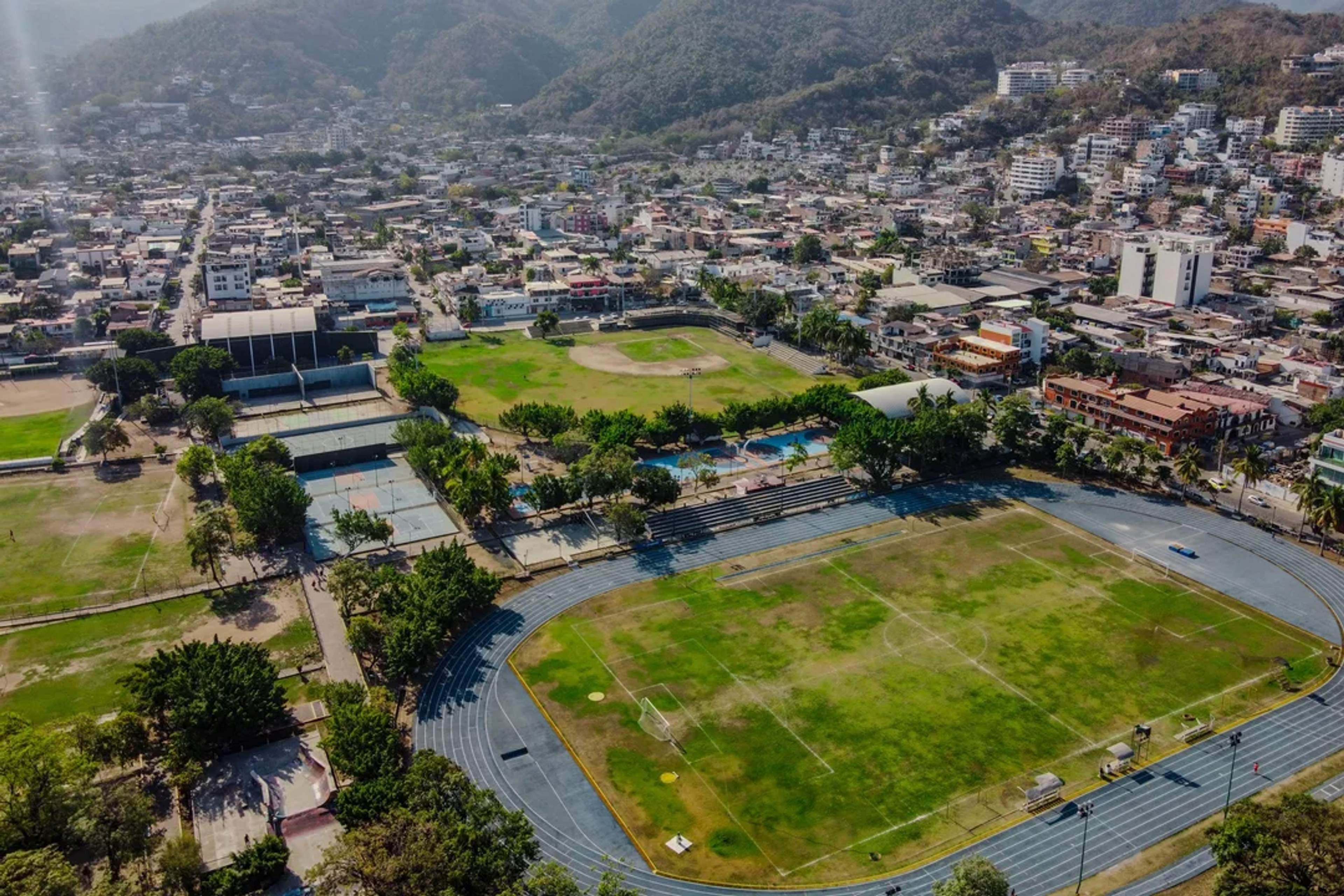 Aerial view of sports complex with soccer field, pool, and courts in a mountainous city.