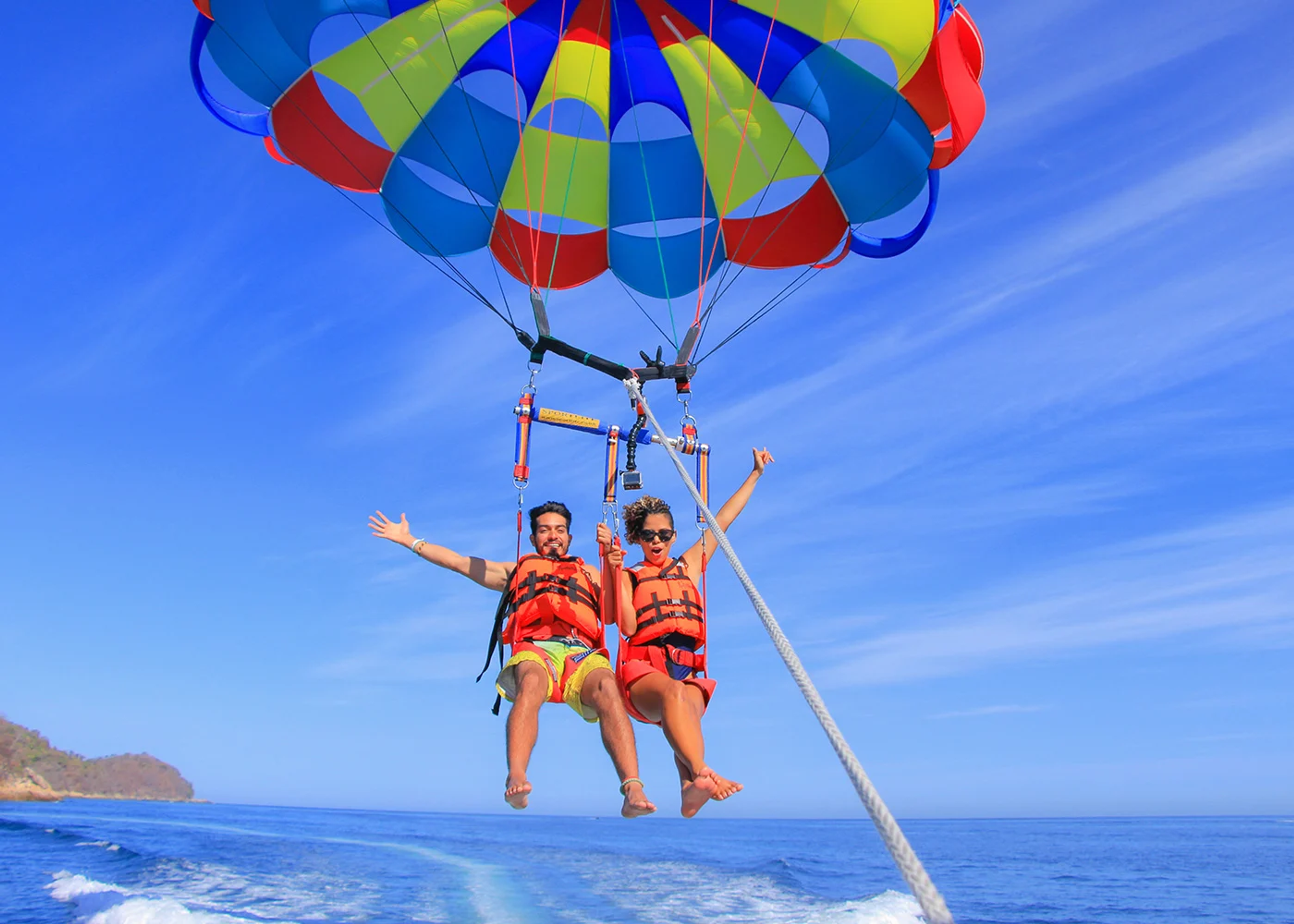 Una pareja disfrutando de una aventura de parasailing sobre el océano, usando chalecos salvavidas y sonriendo con los brazos extendidos. Están atados a un paracaídas colorido con un cielo azul de fondo.