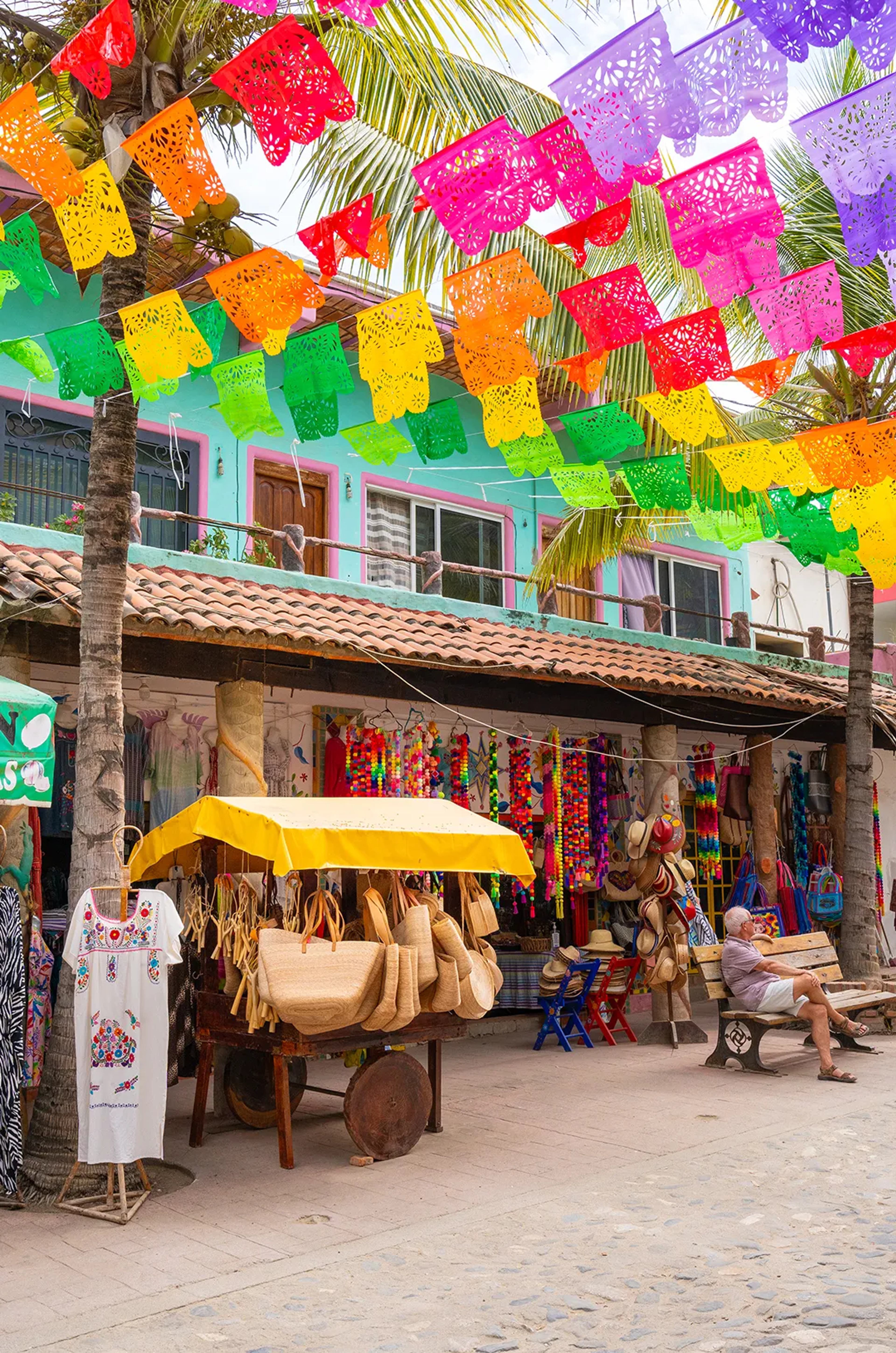 Colorful street market with papel picado, crafts, hats, and a man relaxing on a bench.