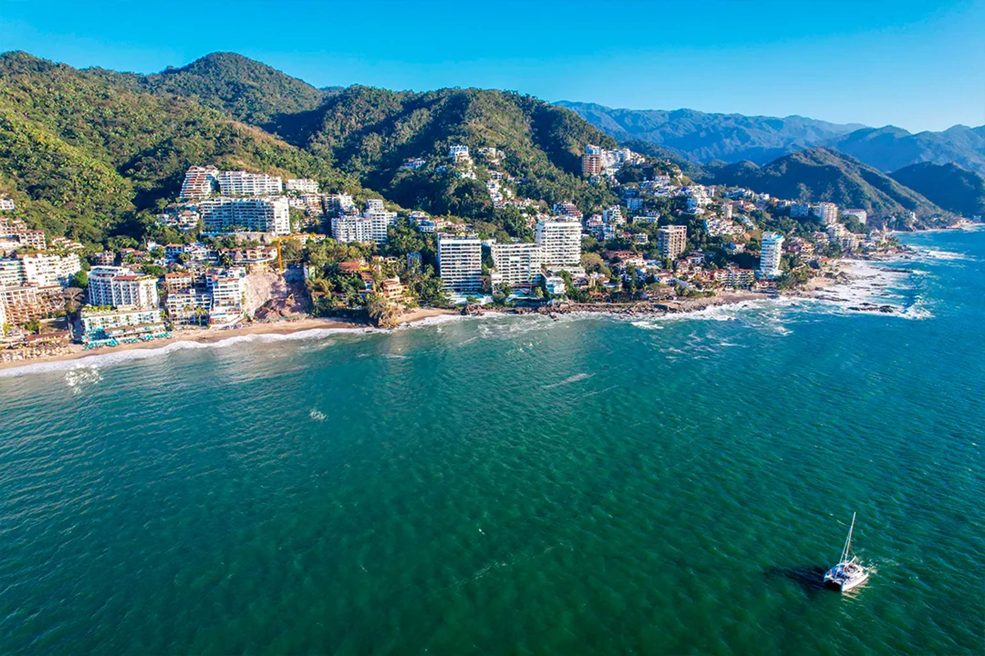 Vista aérea de la costa turquesa de Puerto Vallarta con hoteles frente al mar, acantilados selváticos y botes anclados en la