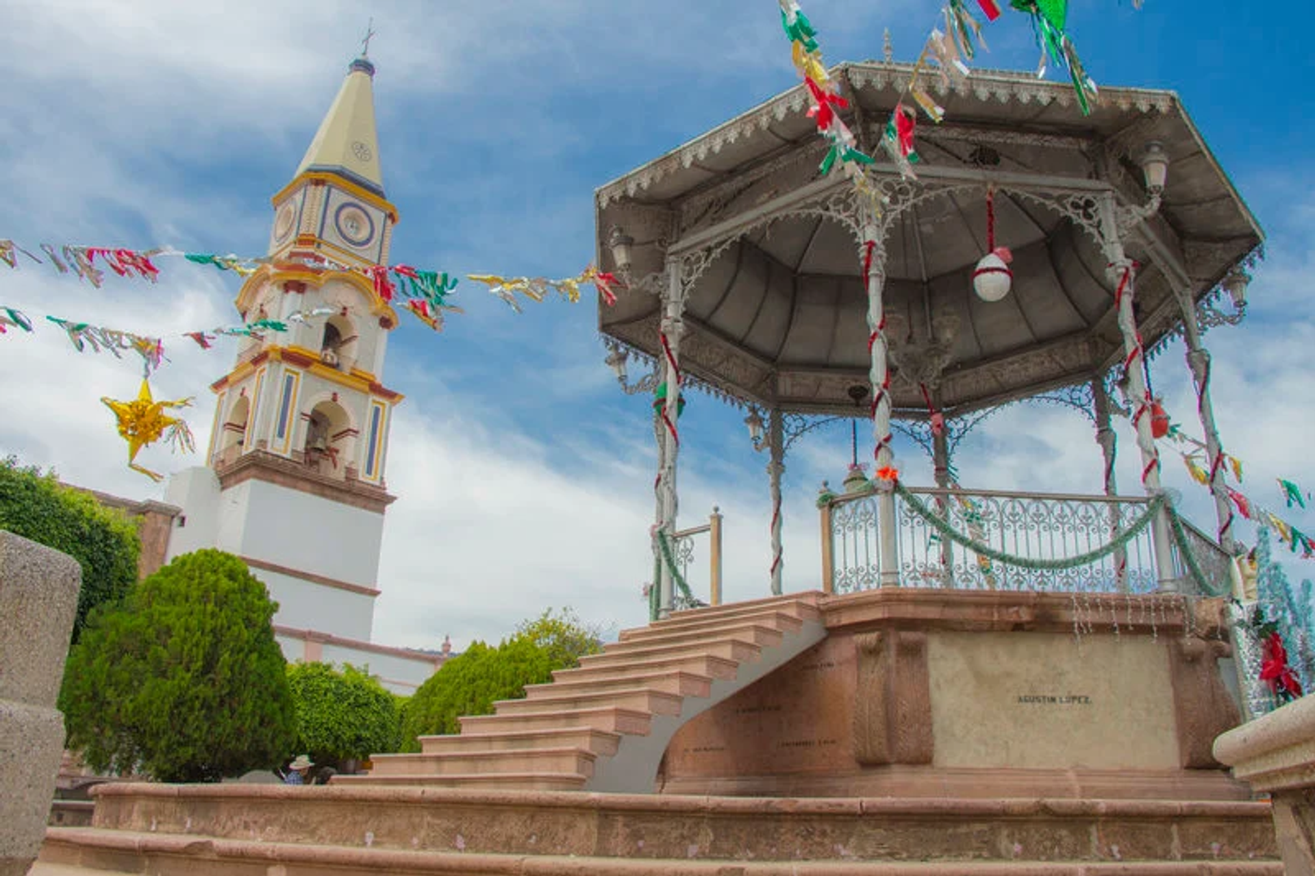 A picturesque gazebo in a Mexican town square, adorned with colorful decorations, with a church tower in the background.