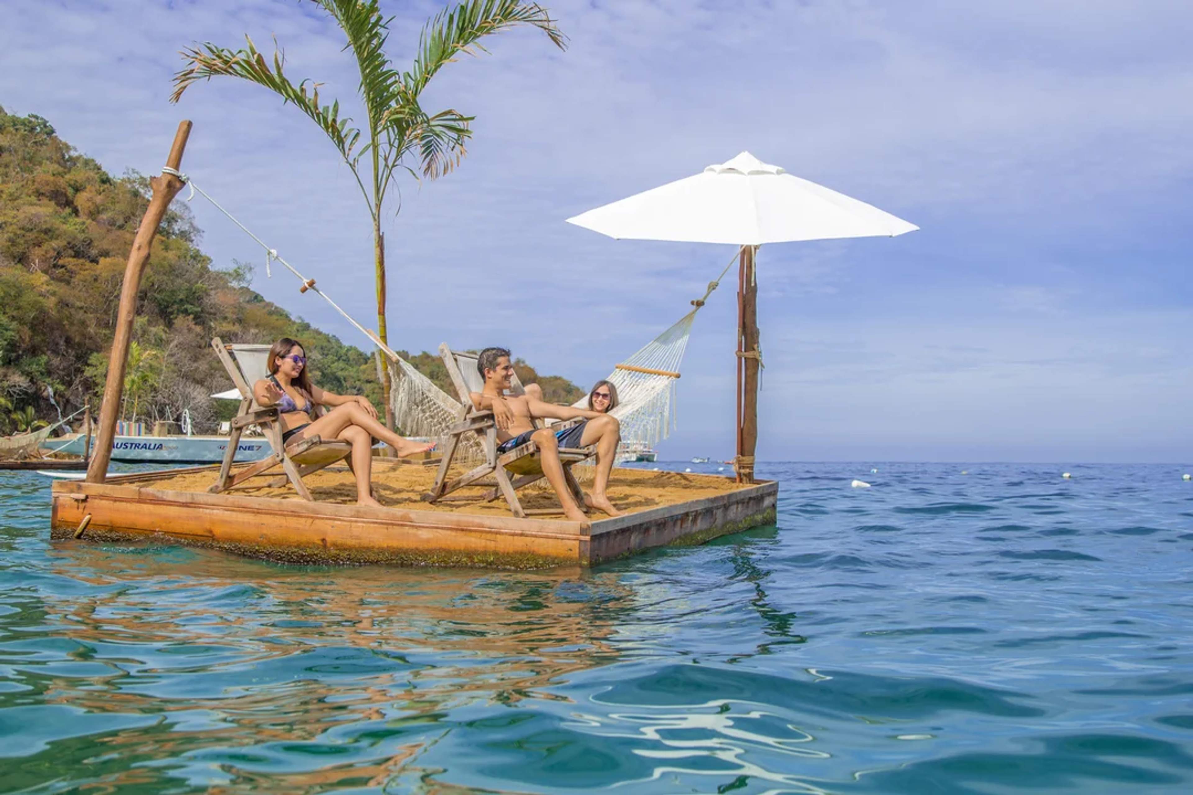 Three friends relax on a floating deck with hammocks, chairs, and a parasol, surrounded by clear blue ocean waters.