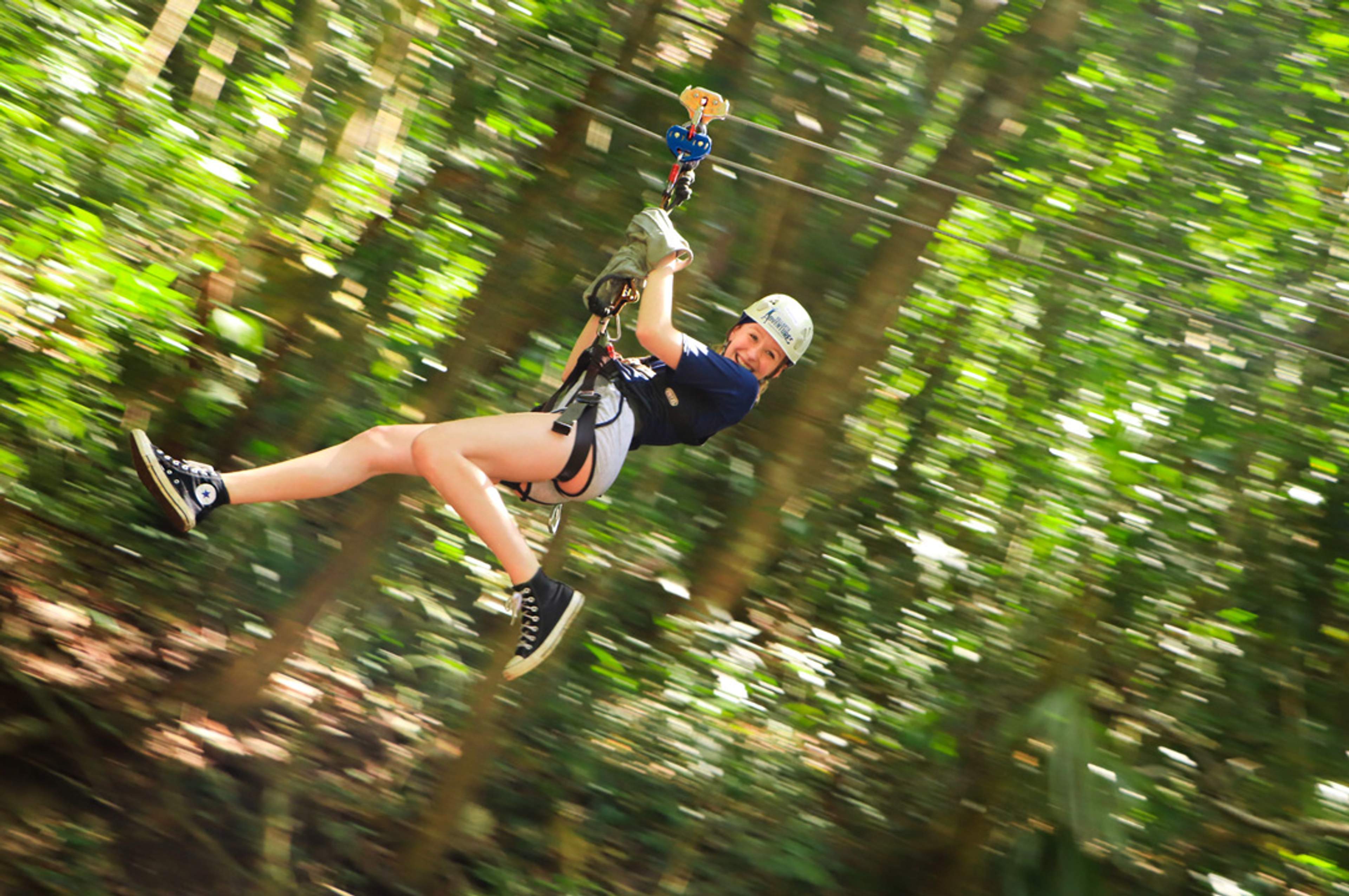 A person wearing a helmet and gloves rides a zipline through a lush forest, smiling and extending one arm out.
