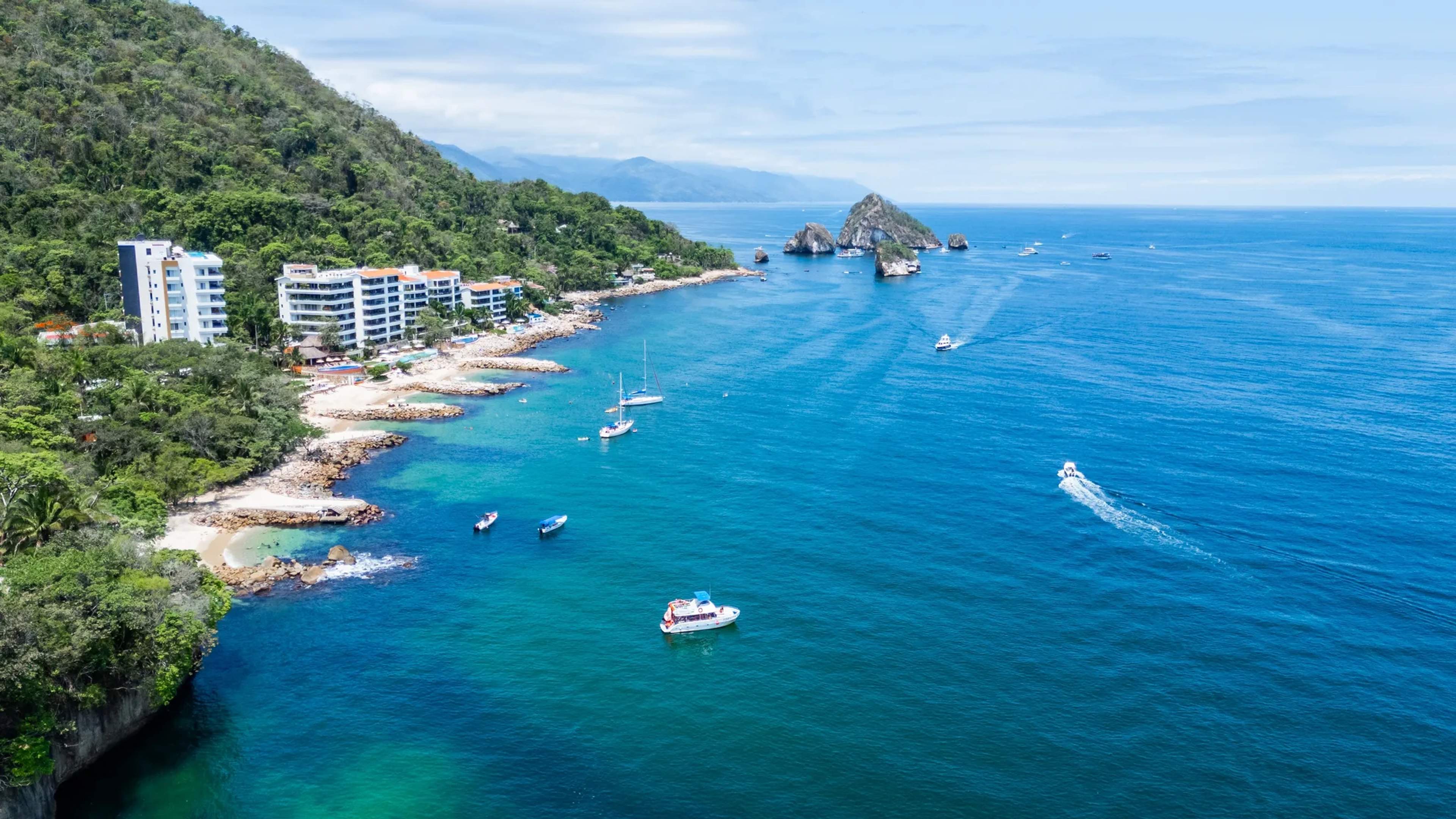 Aerial view of Puerto Vallarta's turquoise coastline, beachfront resorts, jungle cliffs, and boats anchored near rocky