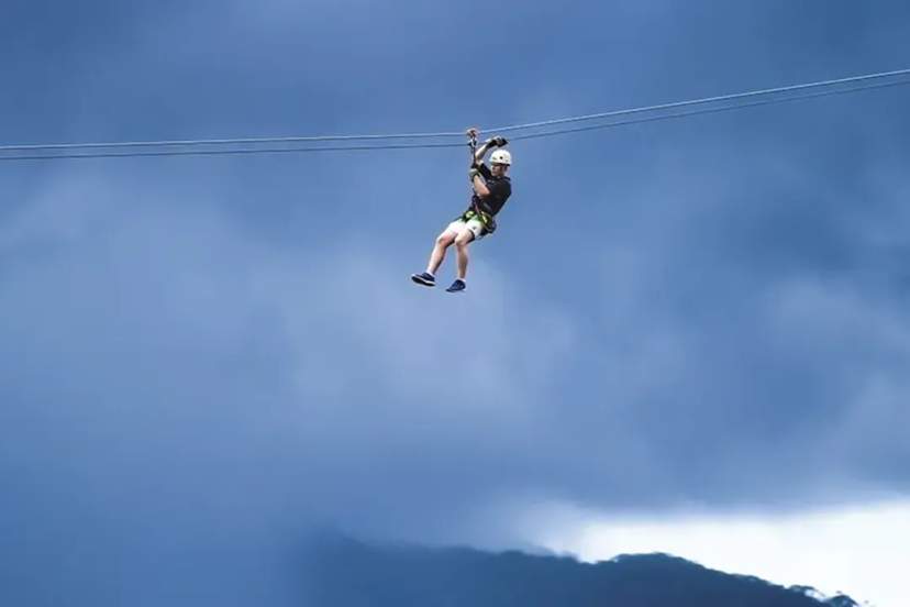 Person zip-lining across the sky with mountains below and dramatic clouds in the background.
