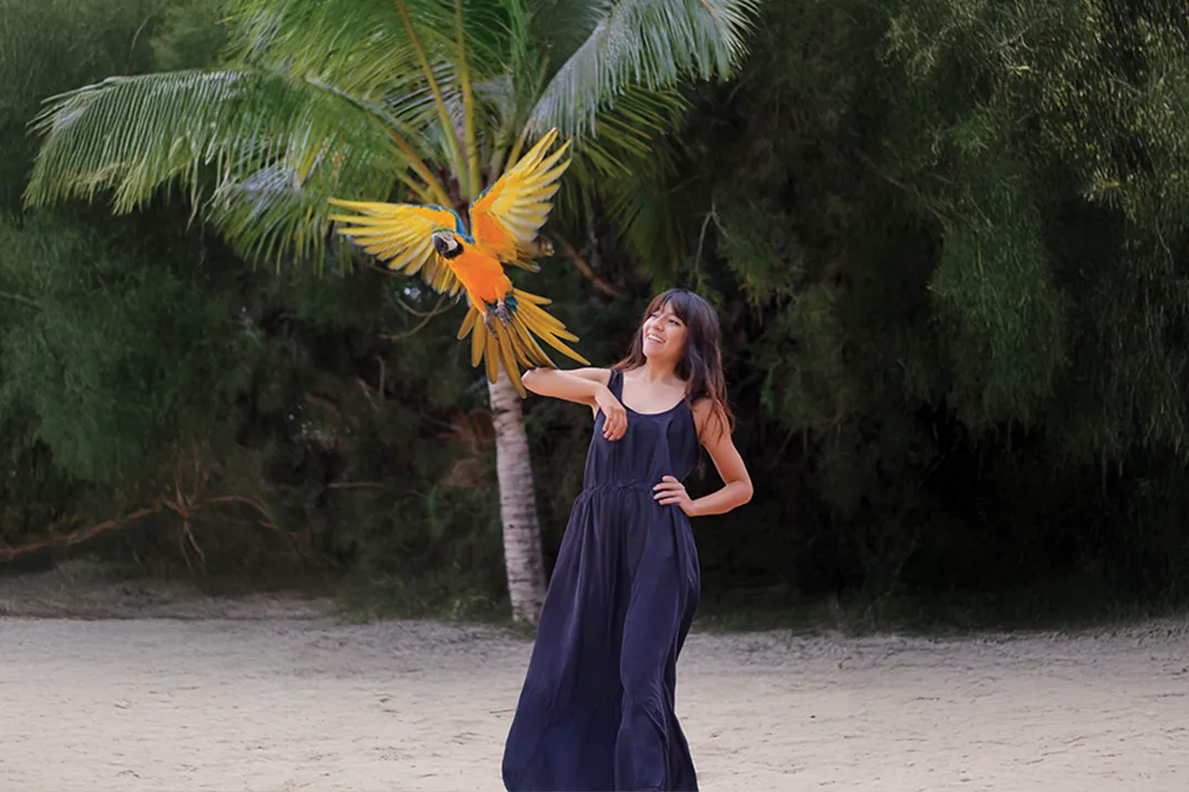 Mujer interactuando con una guacamaya colorida en una playa tropical rodeada de palmeras y naturaleza