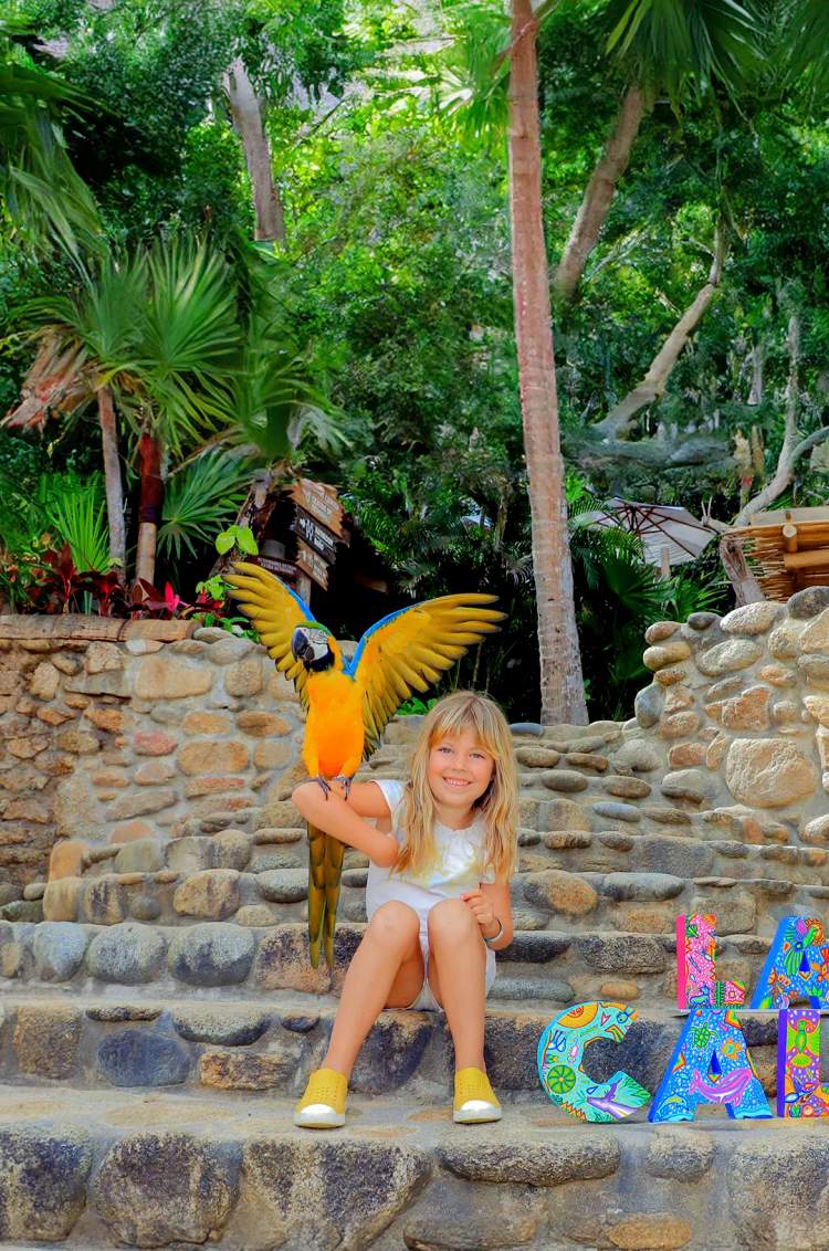 Smiling girl with a macaw on her arm, sitting by "Las Caletas" sign.