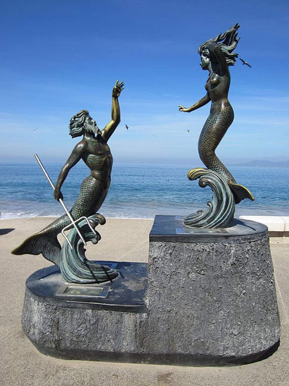 Esculturas de tritón y sirena frente al mar, con tritón alzando el brazo y sirena observando desde una plataforma de piedra.