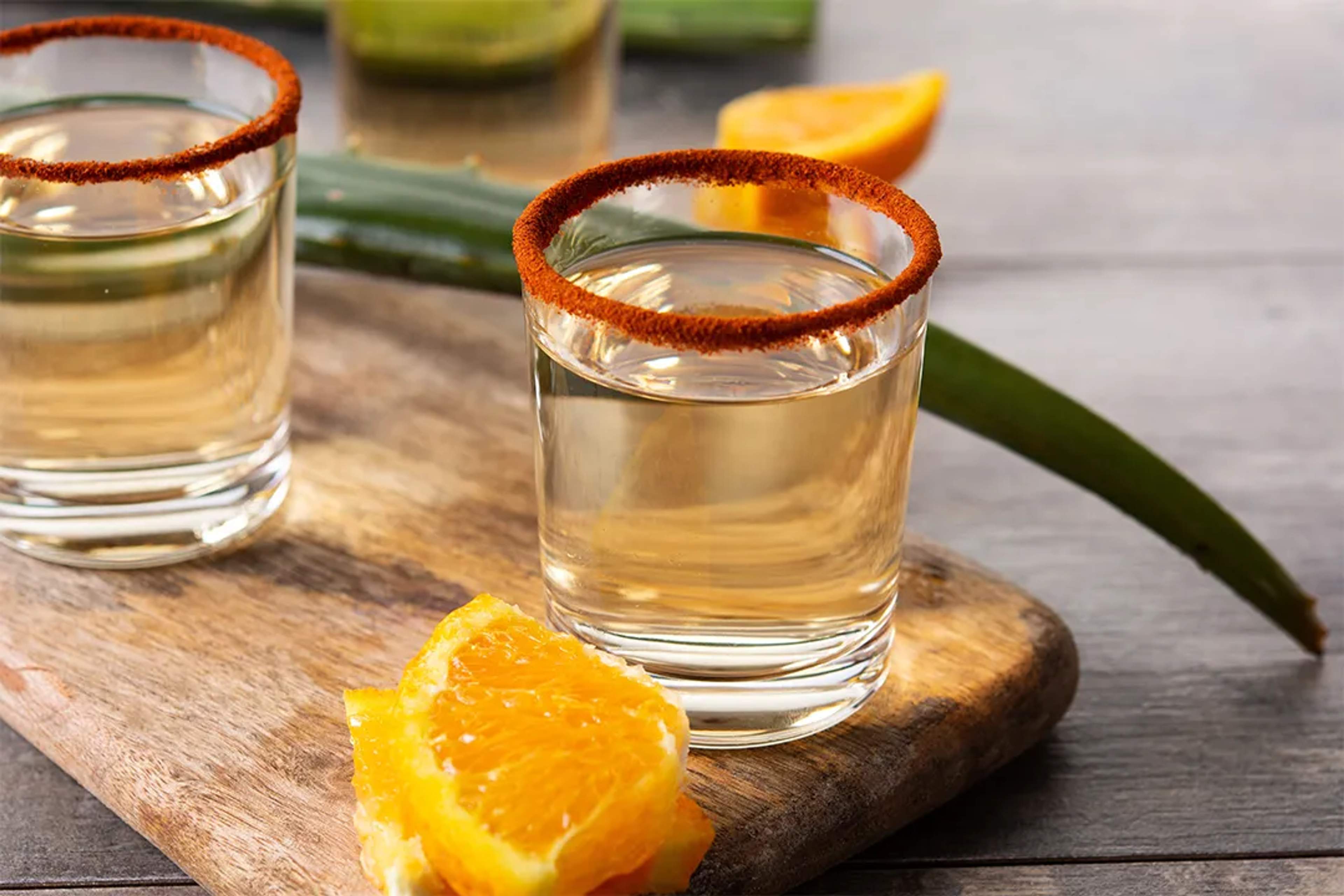 Caballitos de mezcal con chile en el borde y rebanadas de naranja sobre tabla de madera.