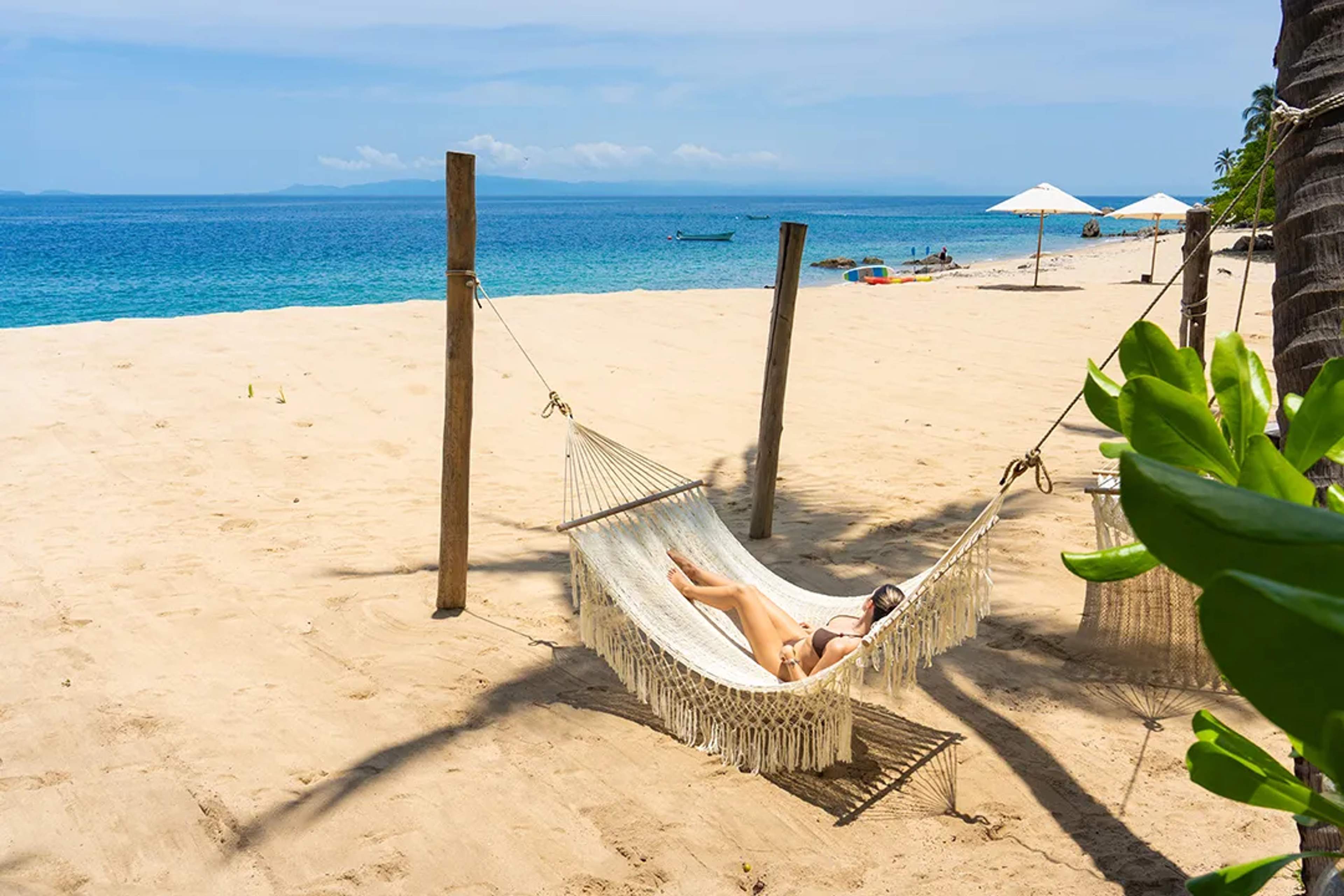 Mujer descansa en una hamaca sobre una playa tranquila de arena dorada y agua turquesa.