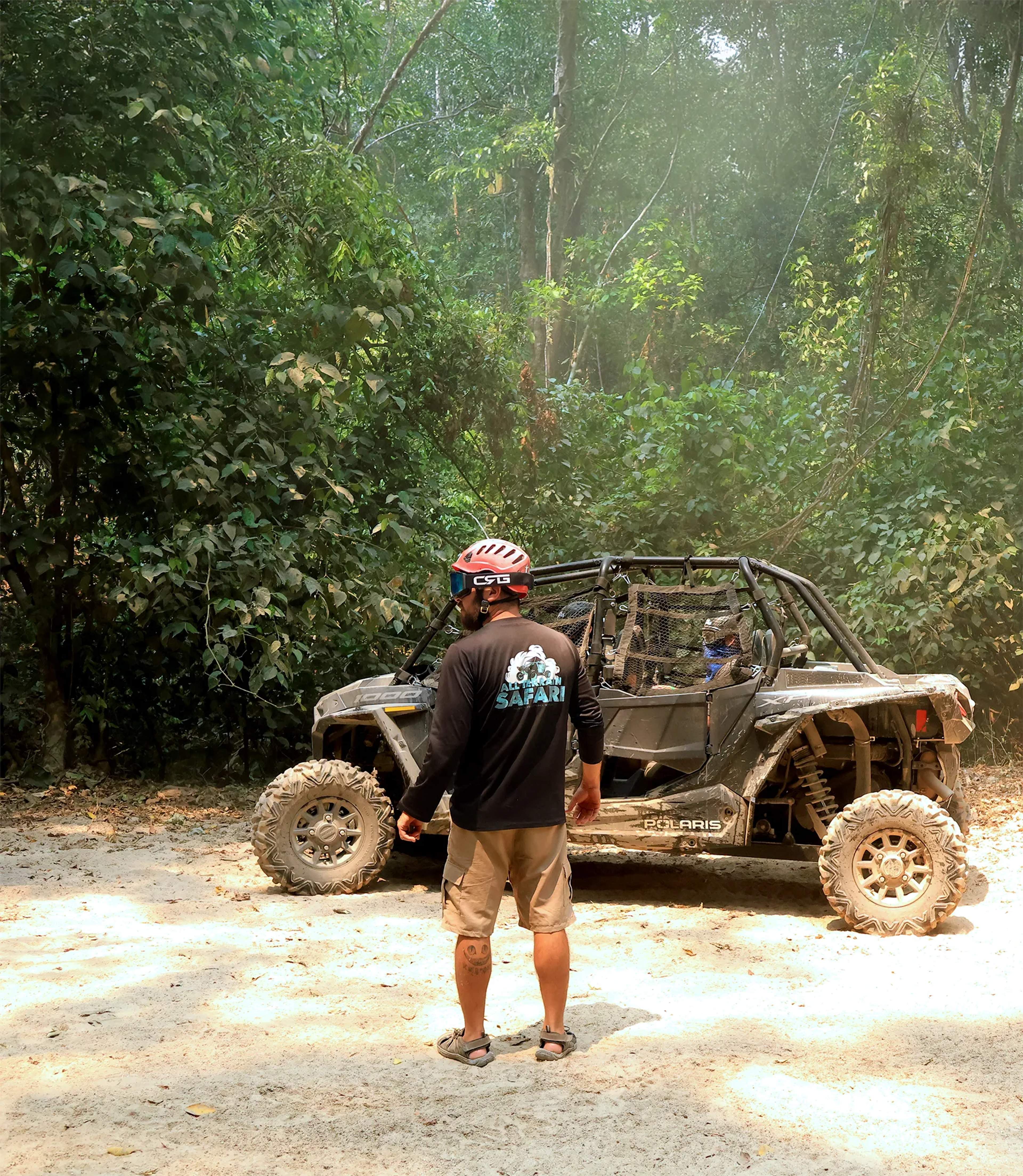 A tour participant stands beside a muddy all-terrain vehicle on a jungle trail surrounded by dense tropical vegetation.