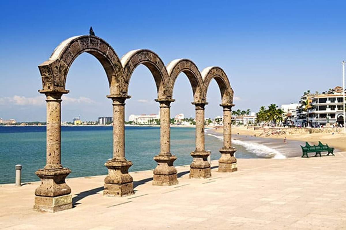 The iconic arches of Puerto Vallarta's Malecón stand against a backdrop of the beach and ocean under a clear blue sky.
