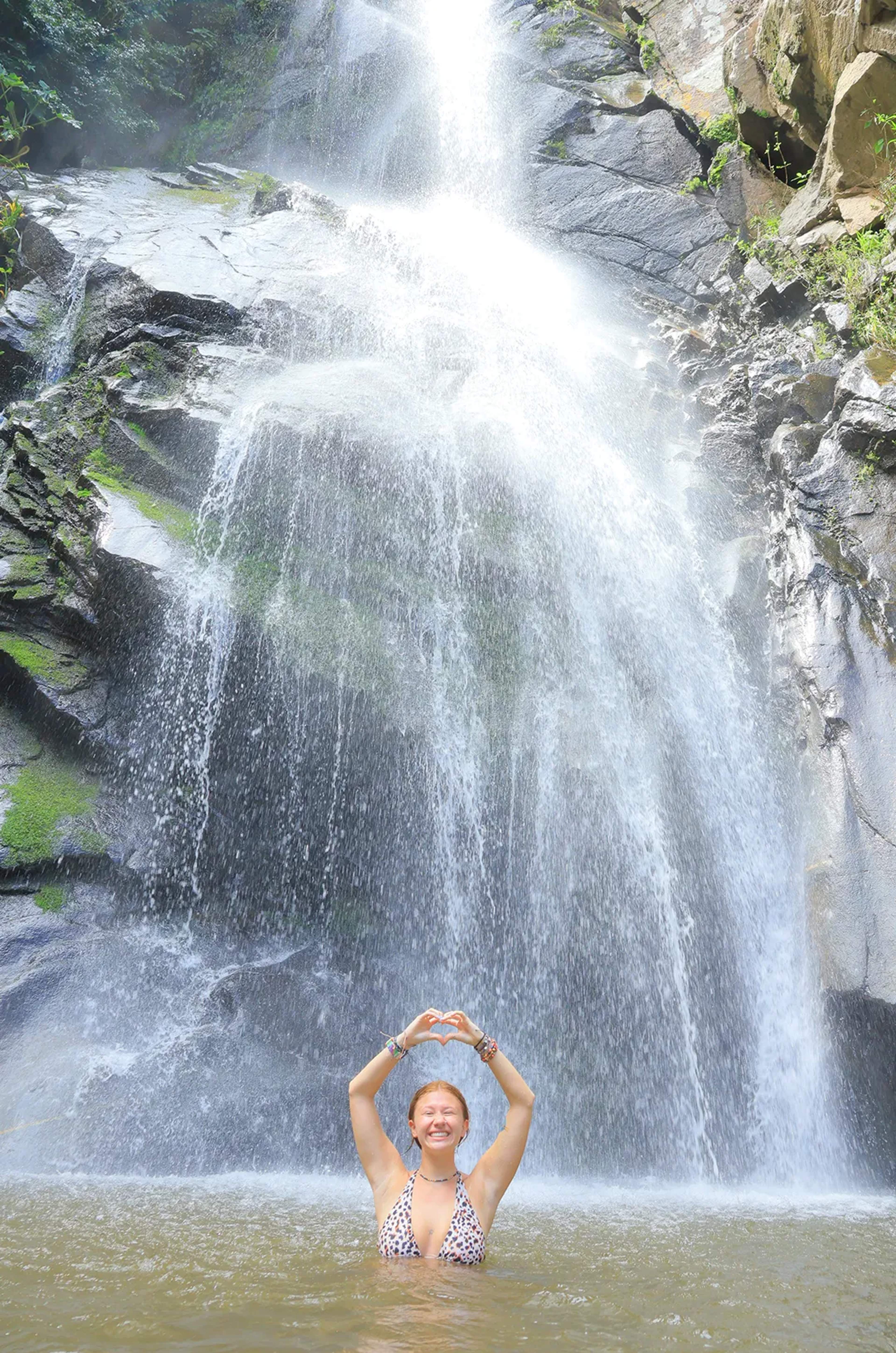 Mujer sonriente en una poza de cascada haciendo un corazón con las manos, rodeada de naturaleza.