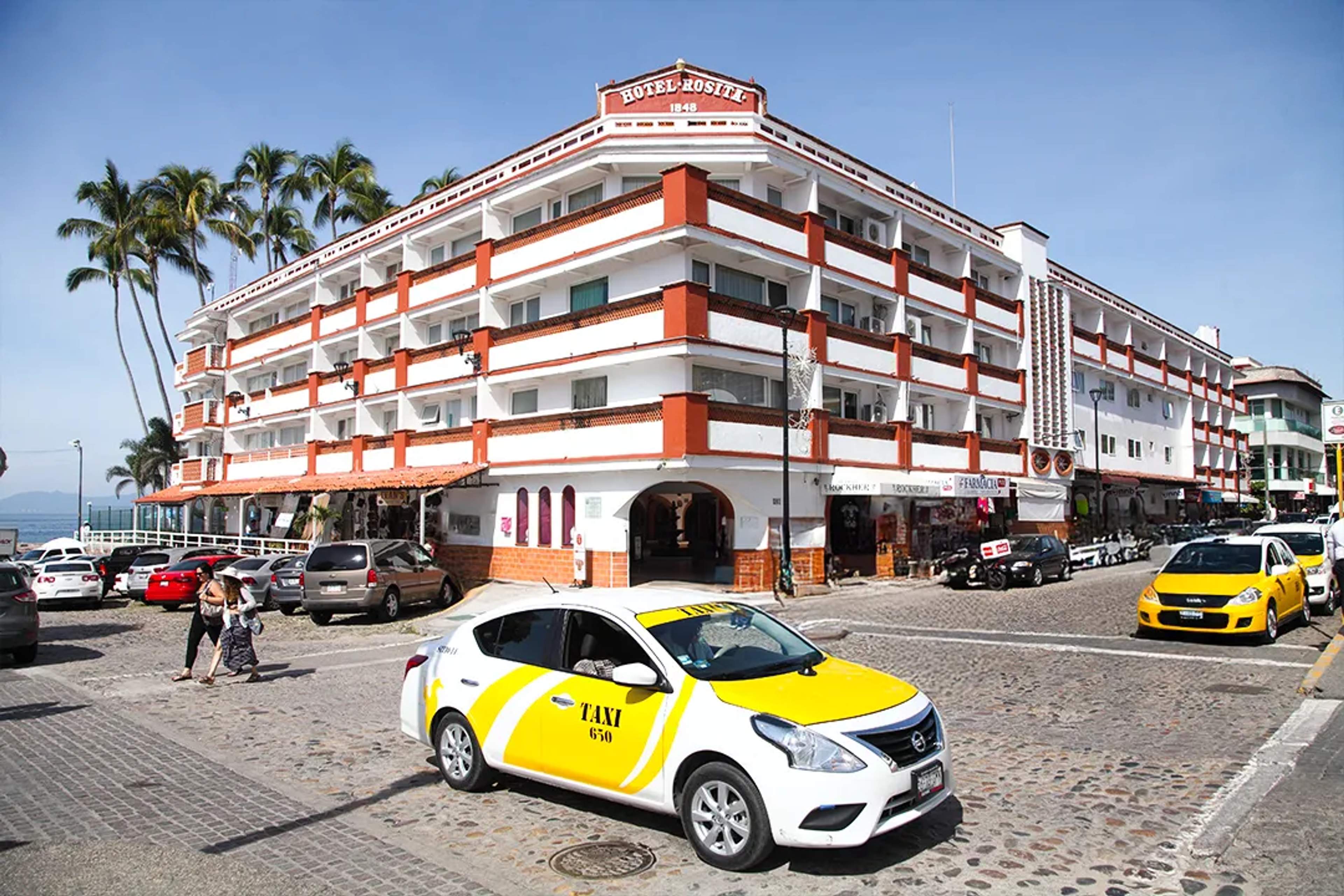 Un taxi pasa junto al Hotel Rosita en el Malecón de Puerto Vallarta, con vista al mar y palmeras en día soleado