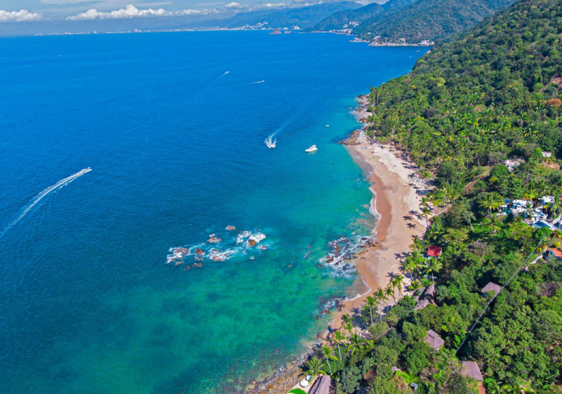 Aerial view of a tropical coastline with lush green forests meeting a sandy beach. The clear blue waters of the bay are dotted with boats and surrounded by rugged hills in the distance.