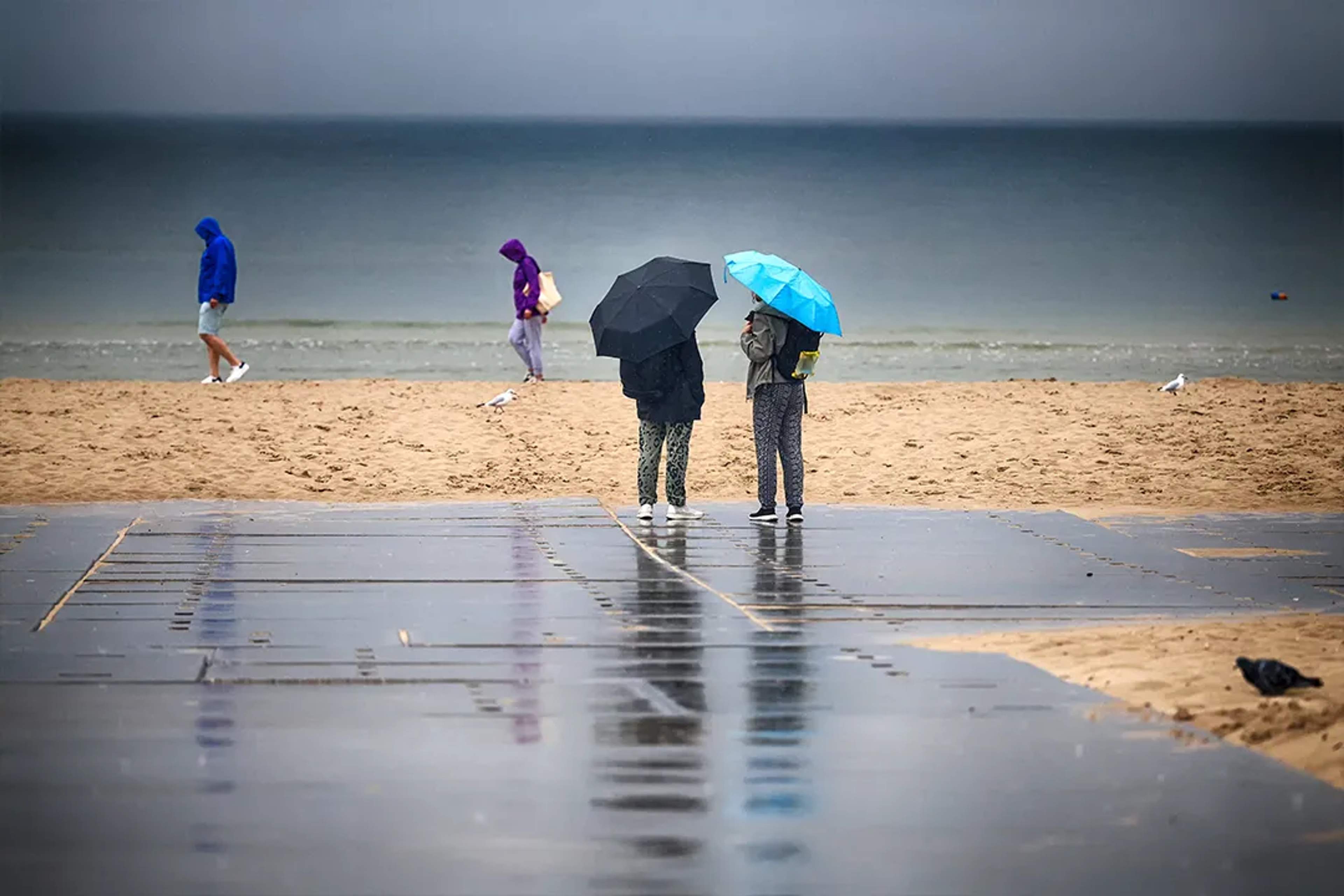 People walk on a rainy beach, wearing hoodies and holding umbrellas under a gray sky.