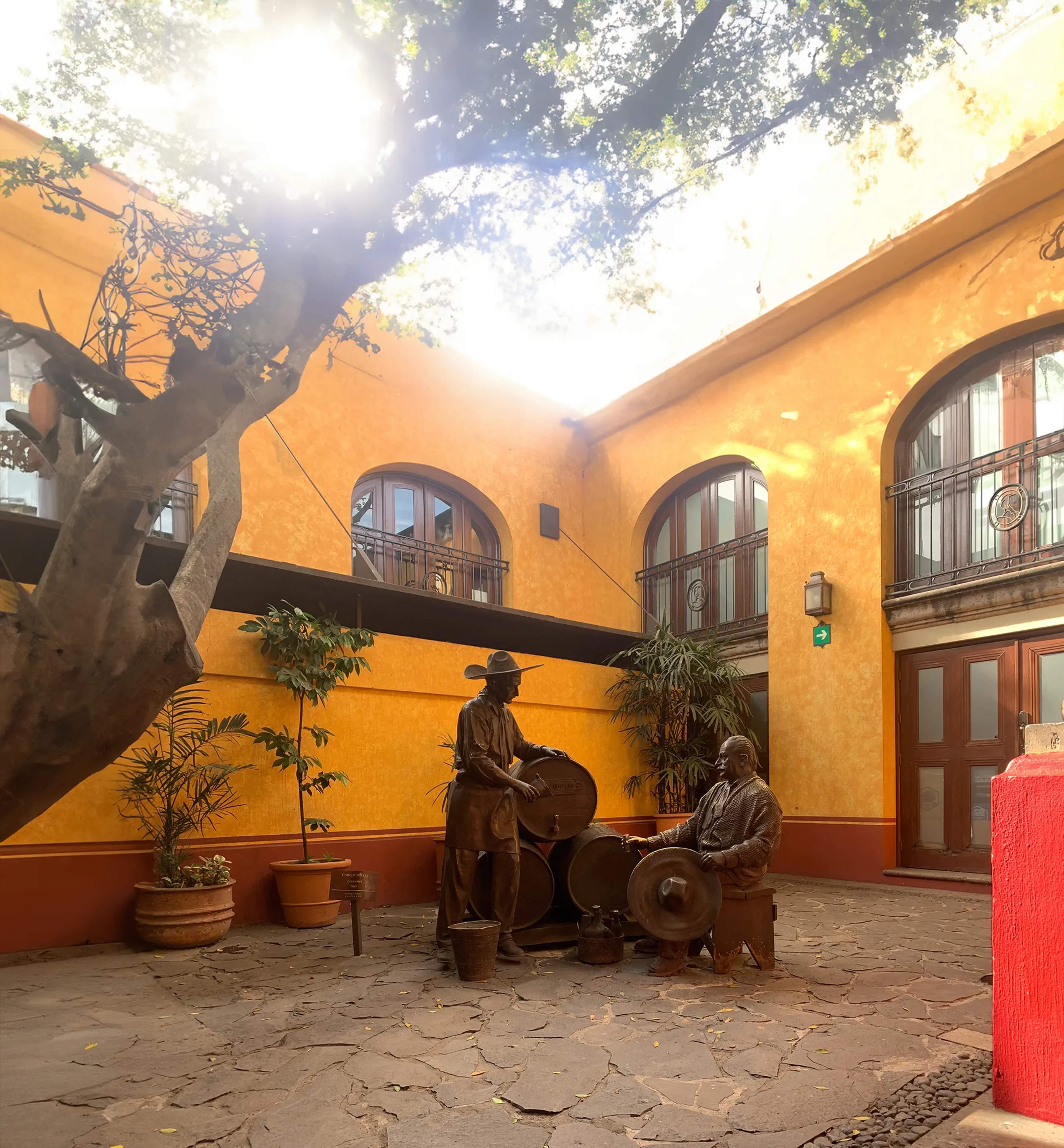 Vibrant colonial courtyard with bronze statues of tequileros, a sprawling tree, and terracotta arches in a Jalisco town