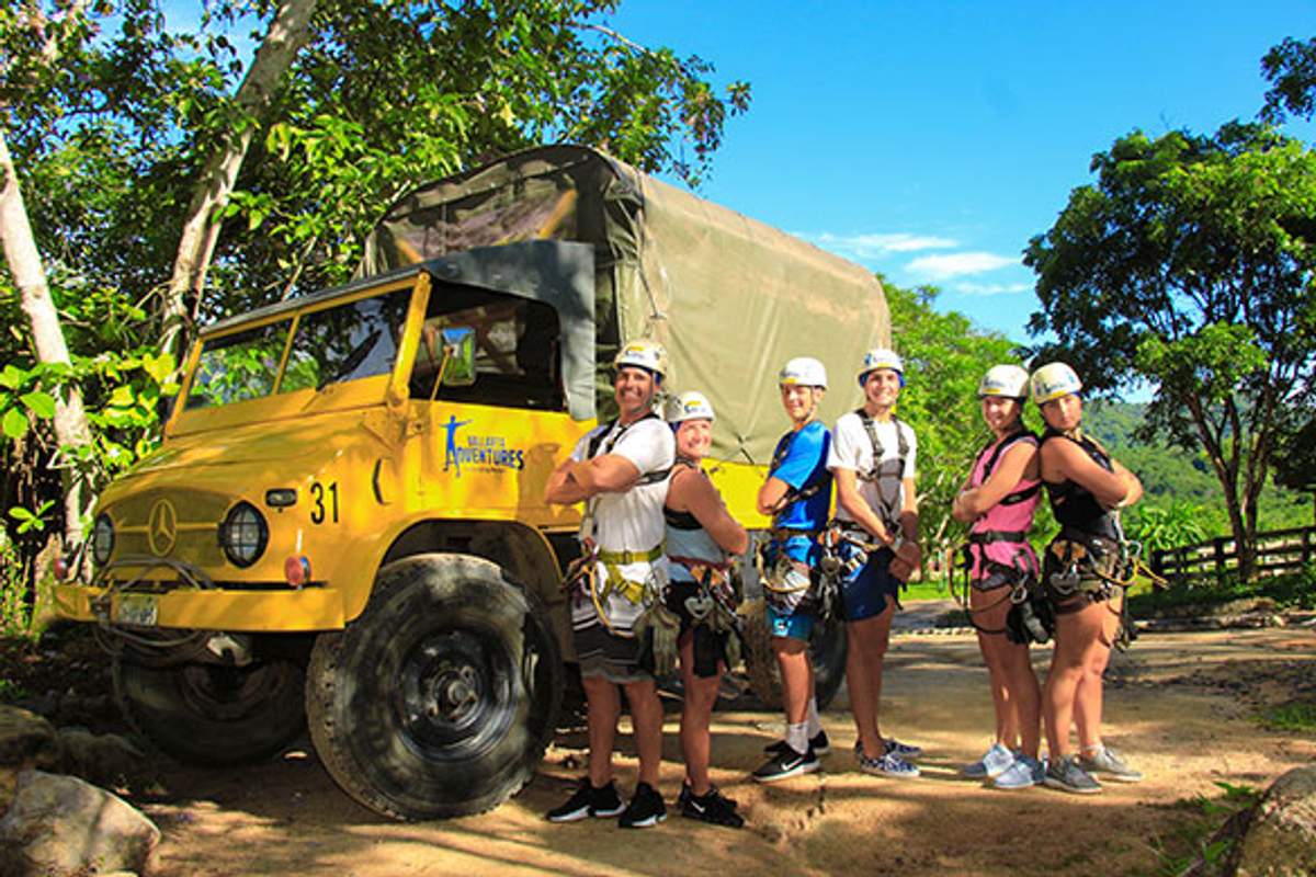 A group of seven people in harnesses and helmets posing beside a yellow adventure tour truck in a lush outdoor setting.