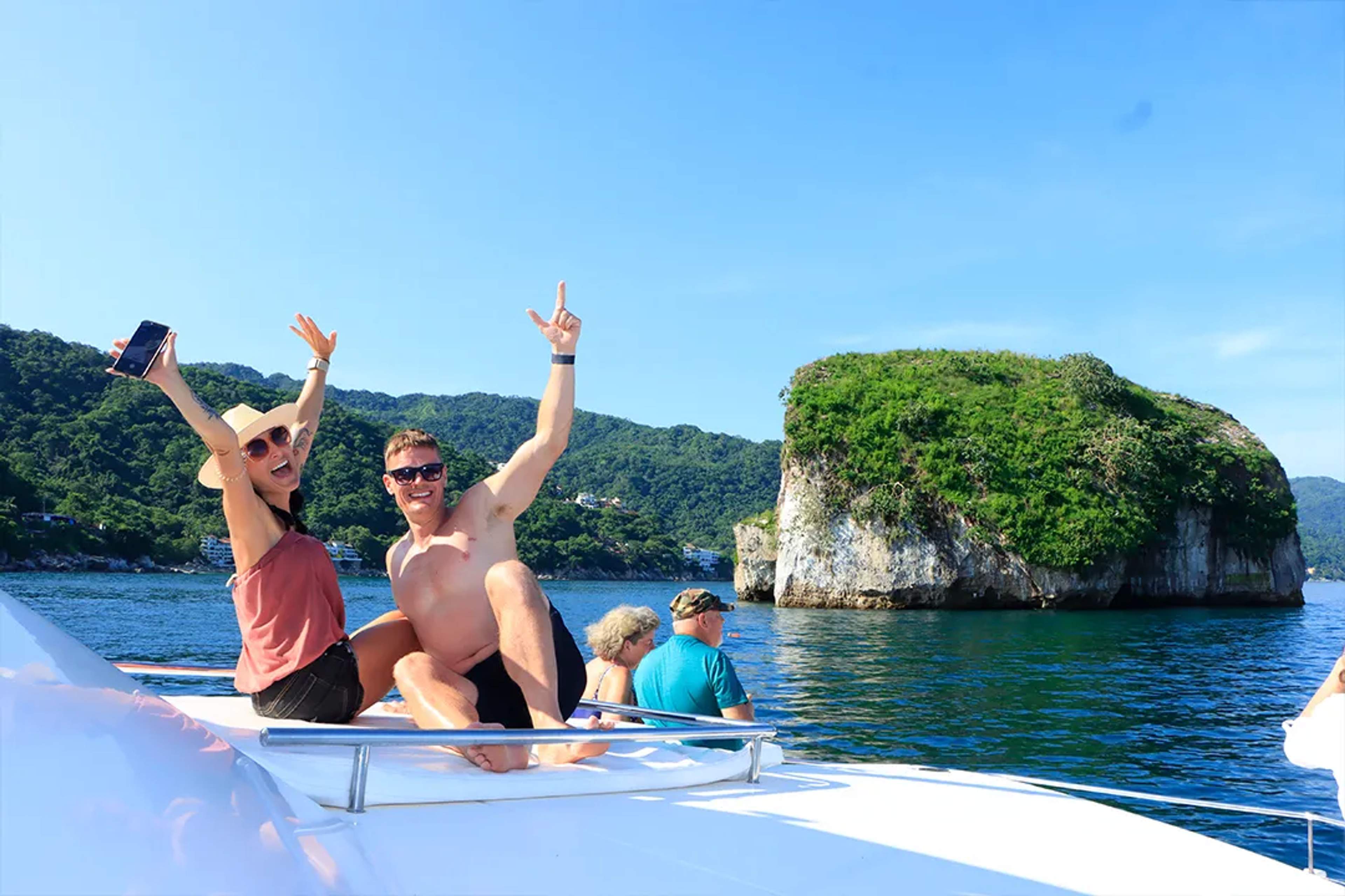 Smiling couple celebrates on a boat with scenic islet and lush mountains in the background.