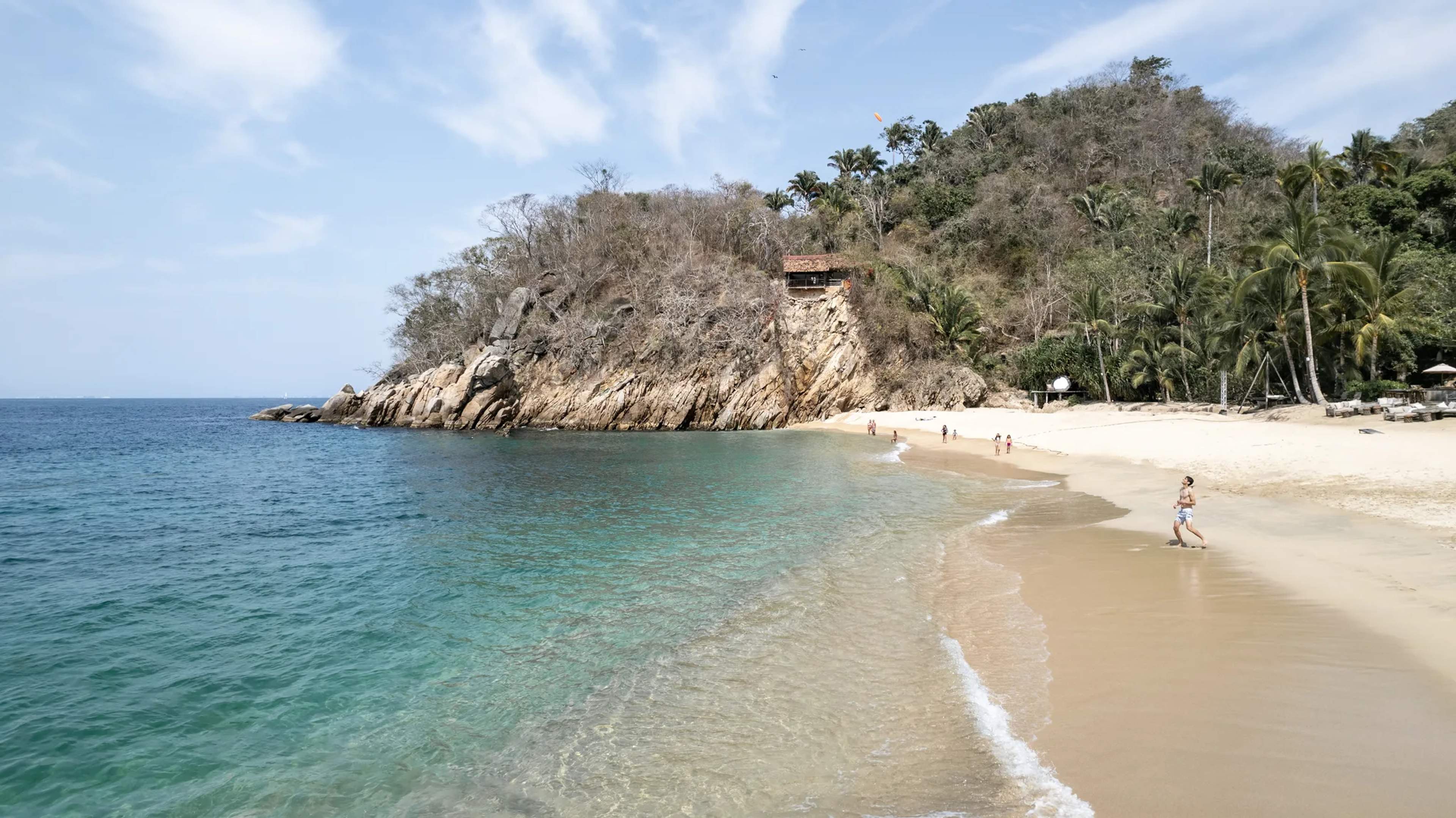 A pristine sandy cove at Majahuitas with turquoise waters, rocky headland, and palm trees, with visitors swimming in calm