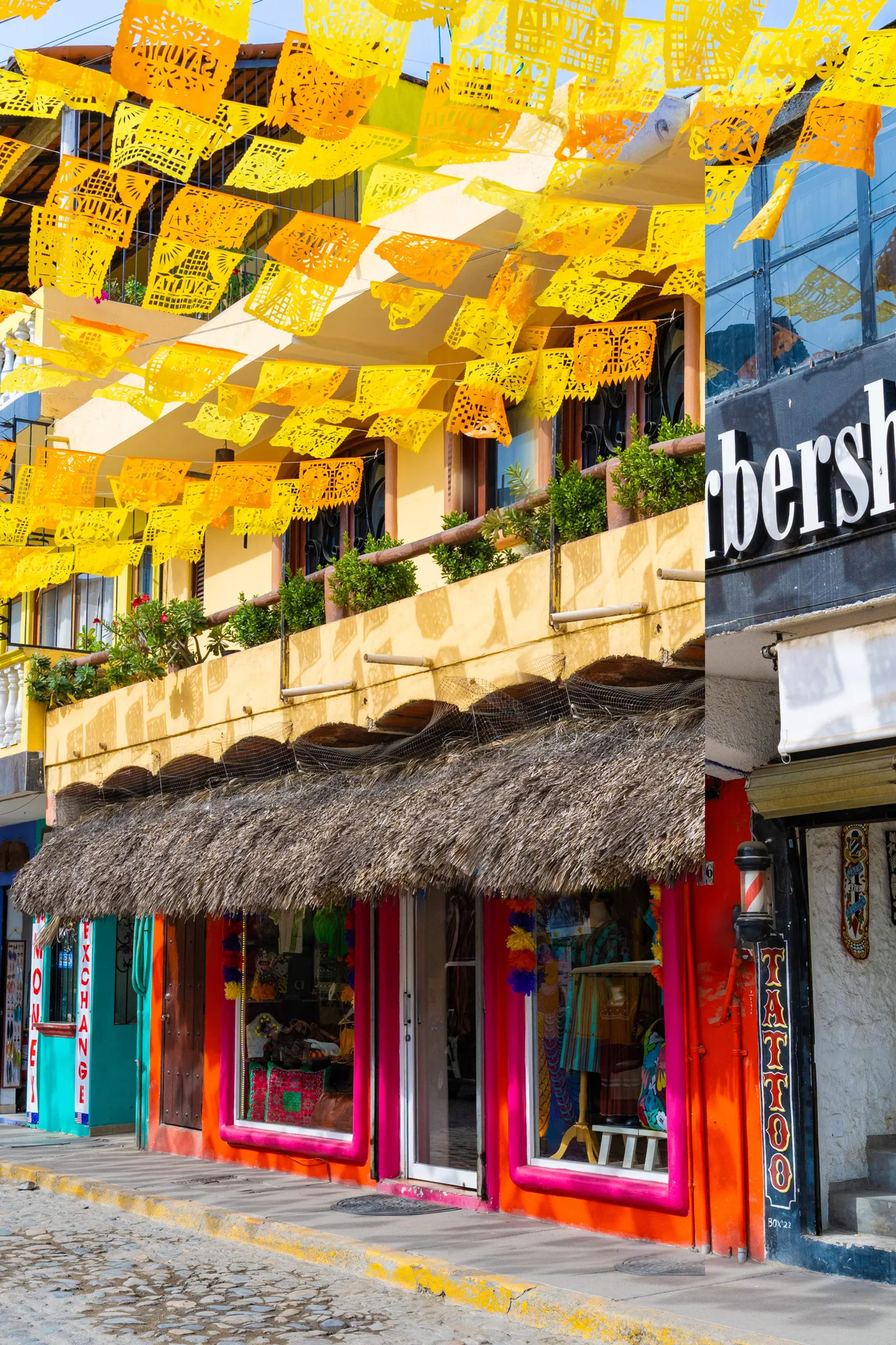 Colorful storefronts on a charming Mexican village street draped with vibrant papel picado bunting overhead.