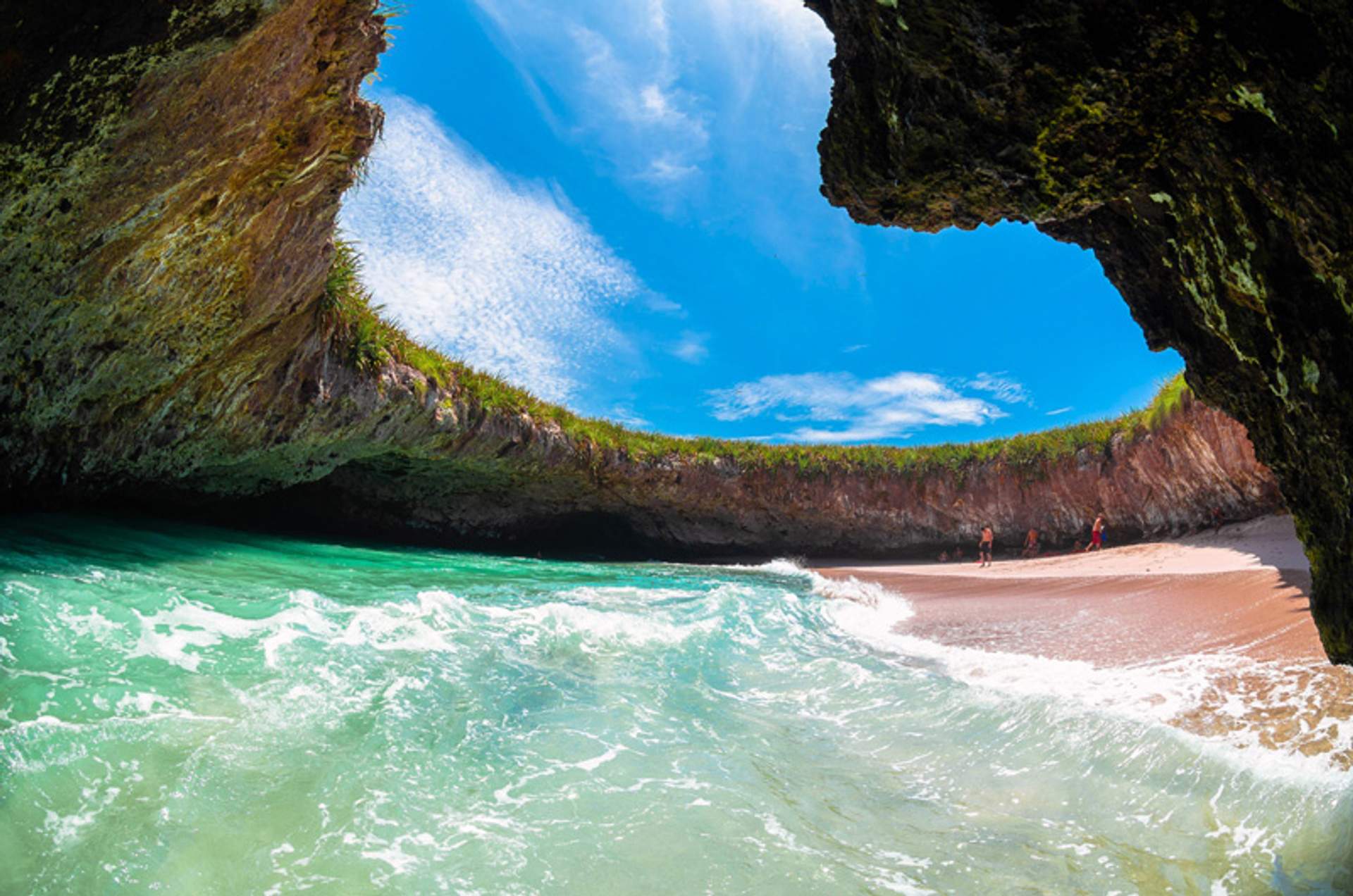 Hidden beach inside a cave with a turquoise sea and sandy shore, under a blue sky.