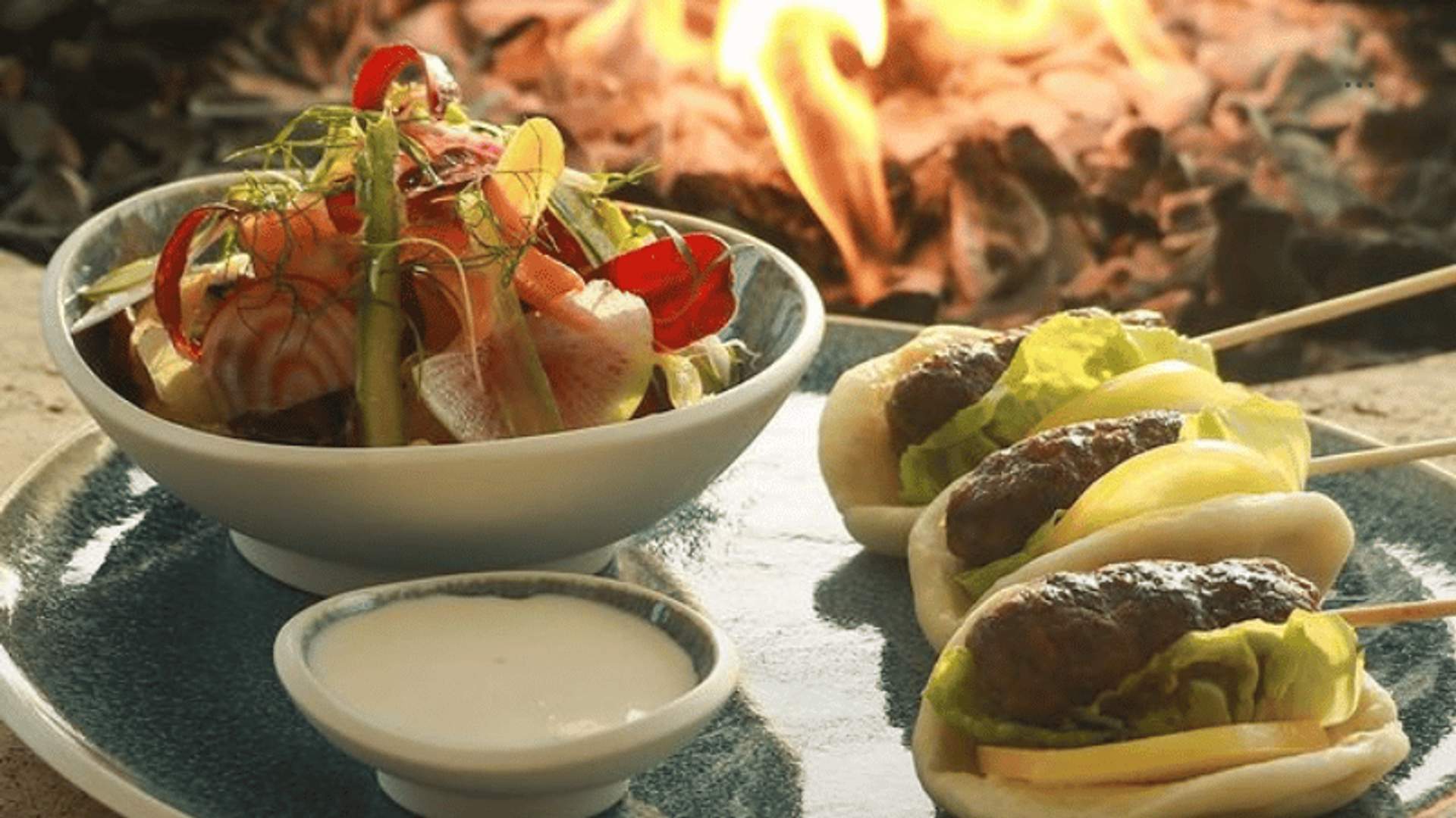 Un plato gourmet que presenta un tazón de ensalada de vegetales frescos y tres pequeños baos rellenos de carne y lechuga, acompañados de una salsa para mojar. El plato está ambientado con el cálido resplandor de una hoguera al fondo.