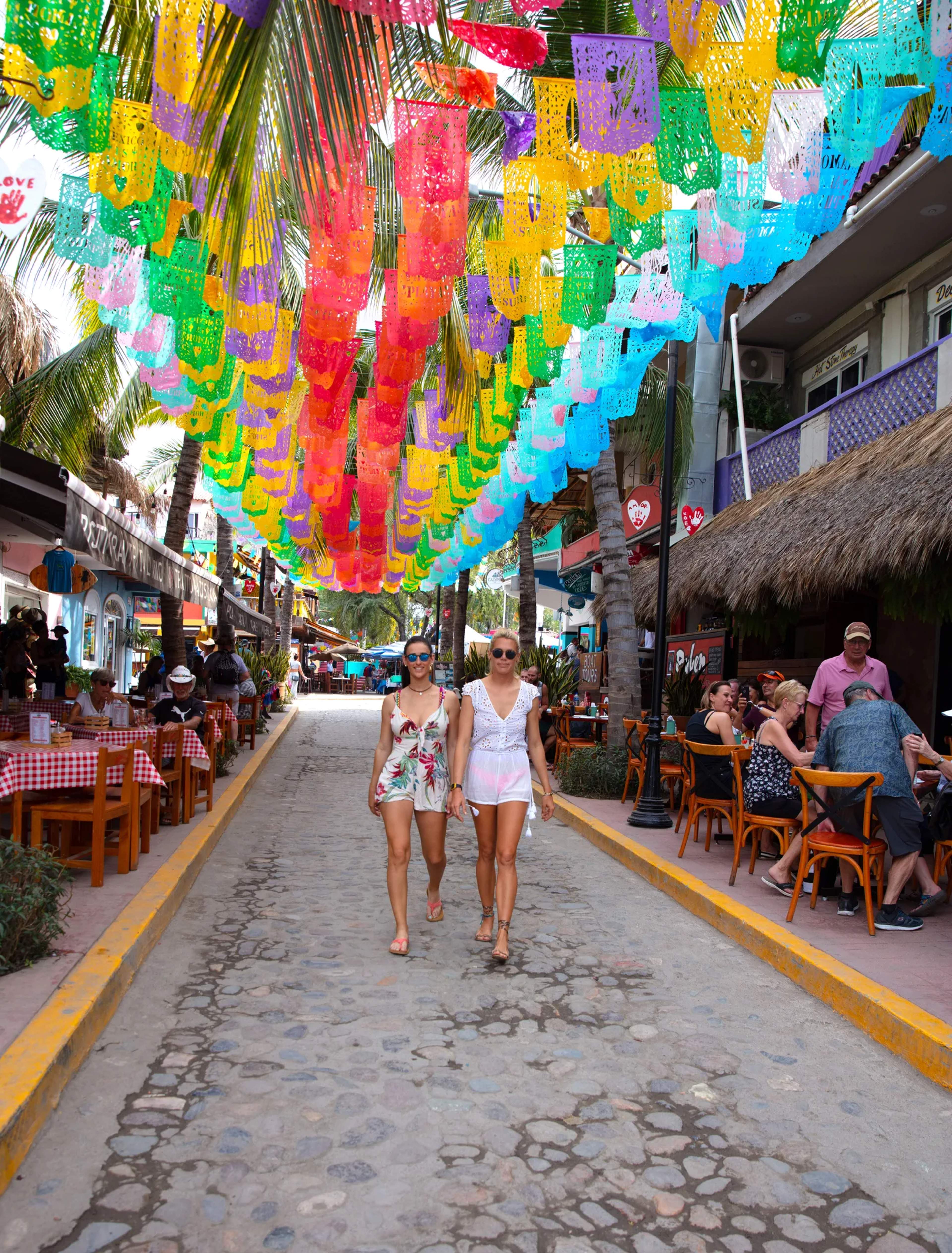 Turistas pasean por una calle de Sayulita adornada con papel picado de colores y hojas de palma.