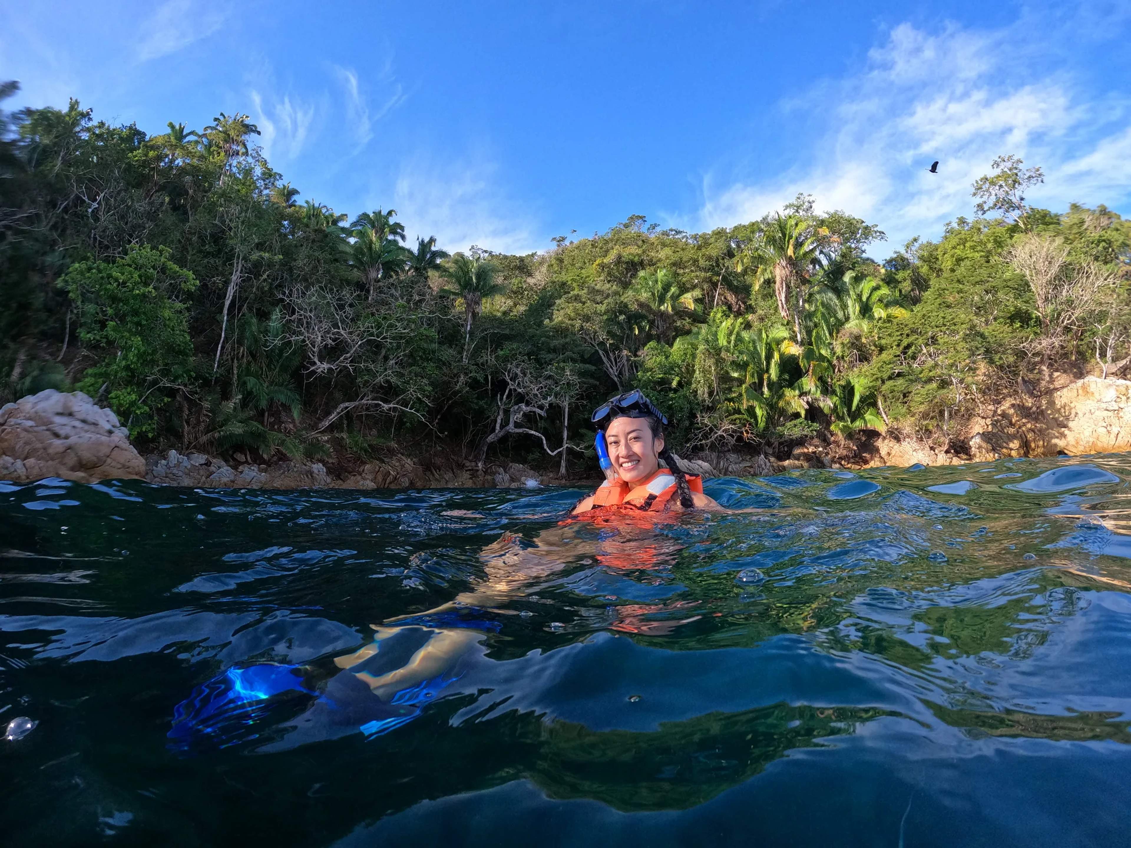 A snorkeler in an orange life jacket smiles while floating in crystal-clear tropical waters near a forested coastline.