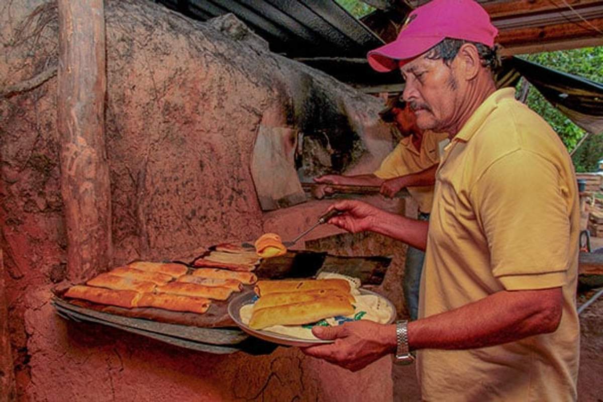 Un hombre con camisa amarilla y gorra rosa saca pan recién horneado de un horno rústico, colocándolo en una bandeja.