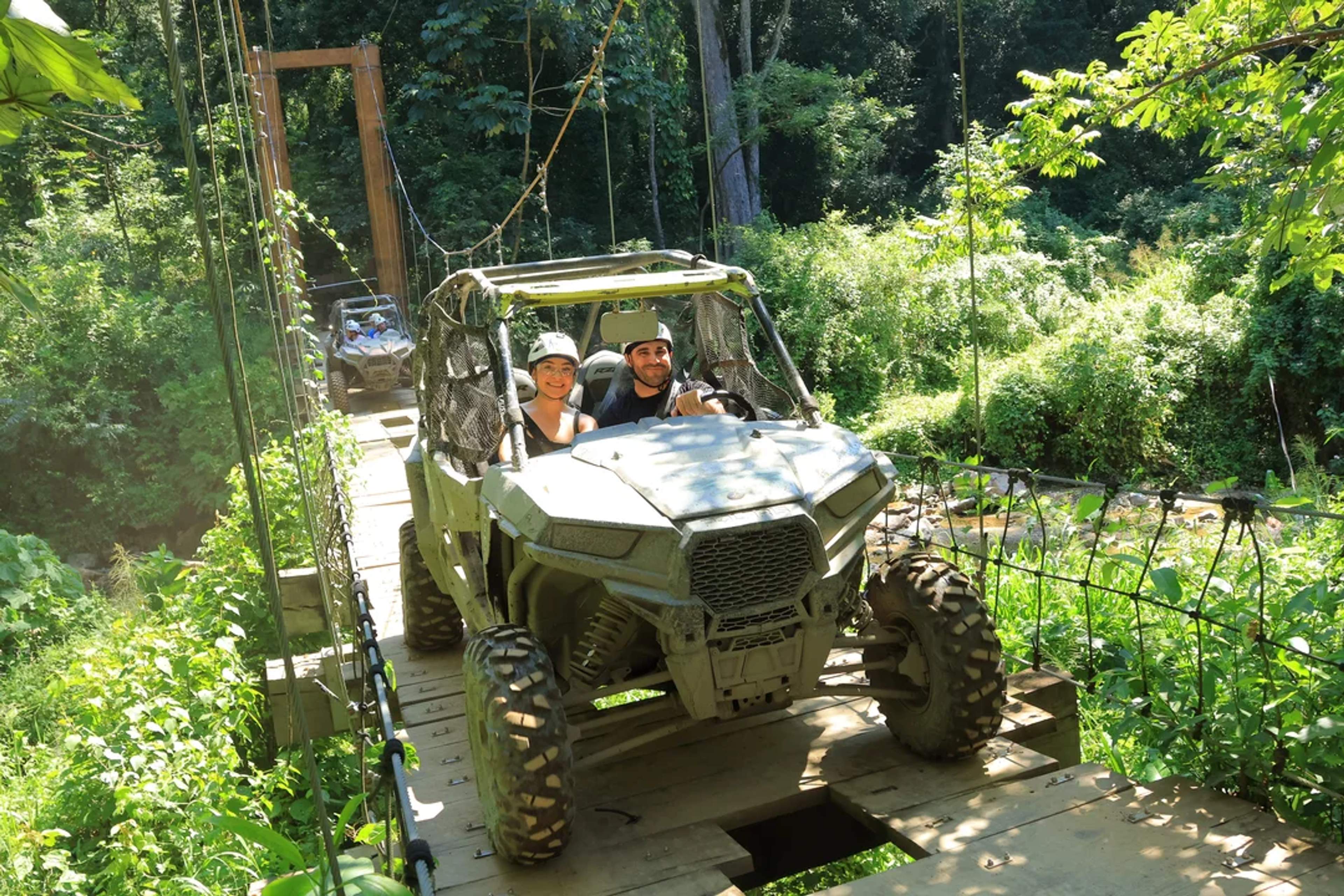 Couple rides a UTV across a wooden hanging bridge surrounded by lush jungle greenery.