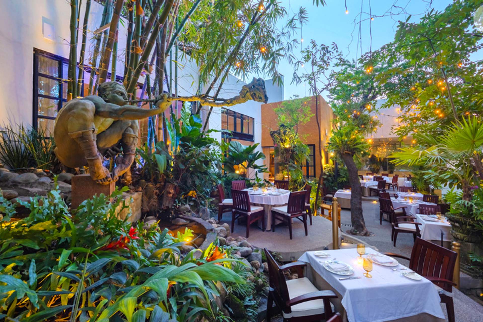An enchanting outdoor dining area at Café des Artistes, adorned with lush greenery, ambient string lights, and a whimsical sculpture.
