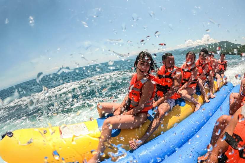 Group of tourists in orange life jackets rides a banana boat off the Mexican coast, waves splashing with mountains in the
